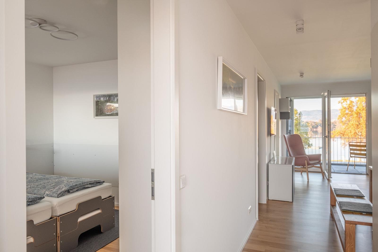 Schlafzimmer mit Bett und Bild. Flur mit Sitzgelegenheit und Balkonblick.