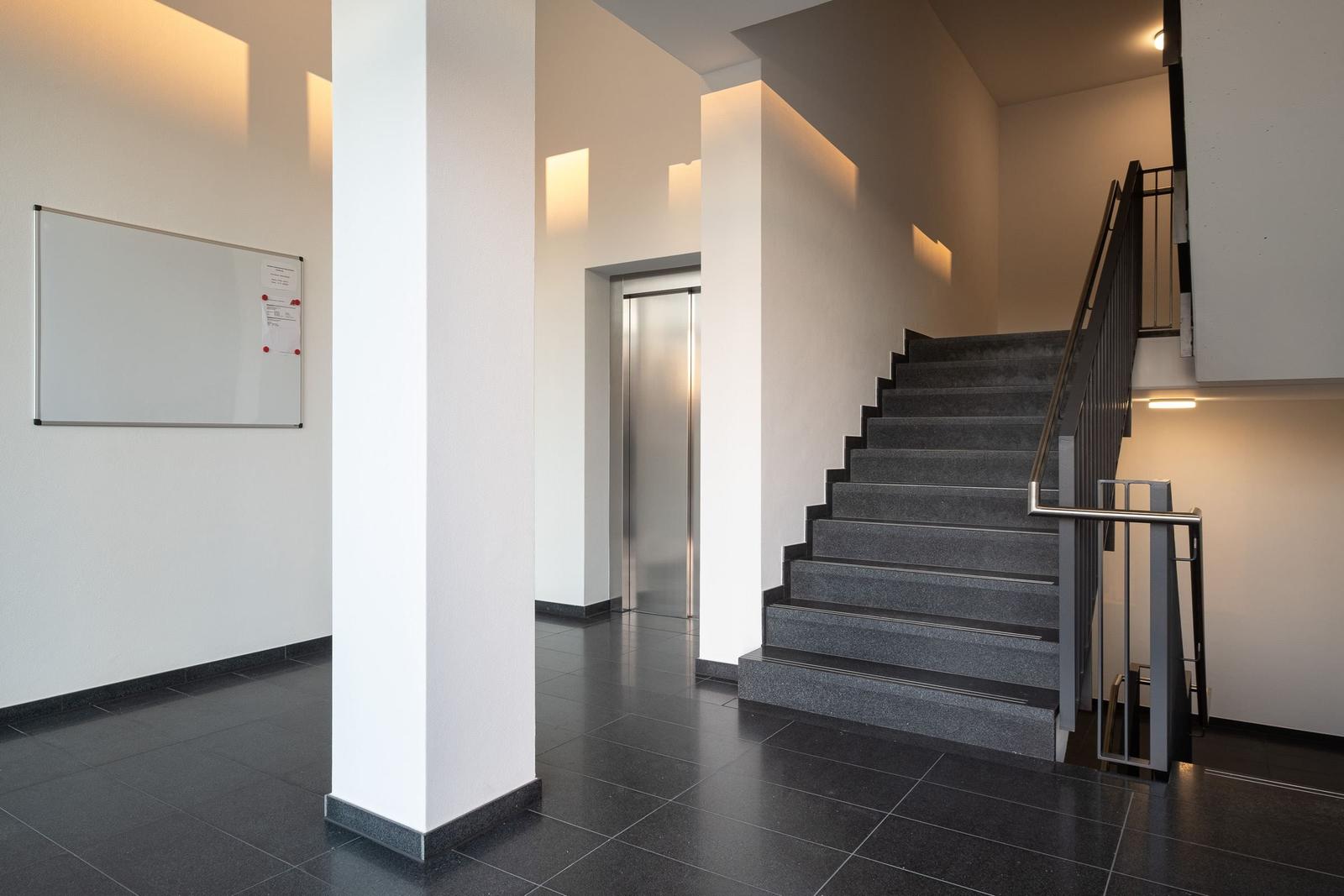 Haupteingang mit Treppe und Aufzug. Weiße Wände und dunkler Boden.