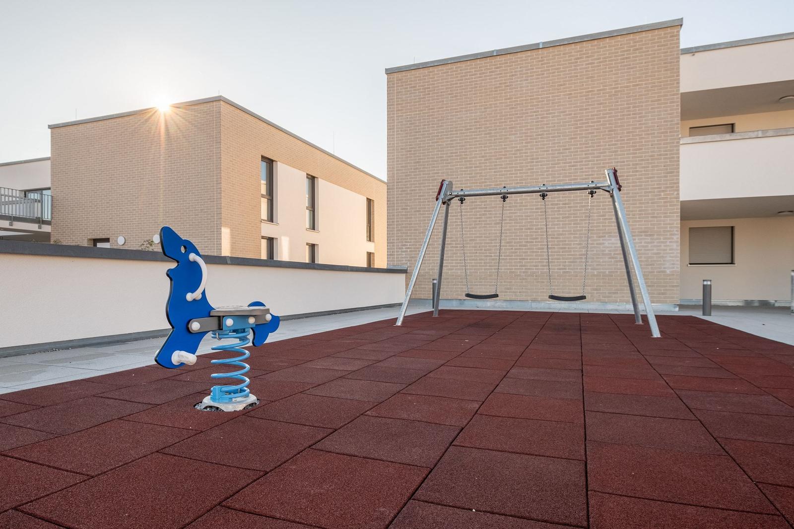 Kinderspielplatz mit Schaukel und blauem Feder-Spielgerät auf rotem Boden.