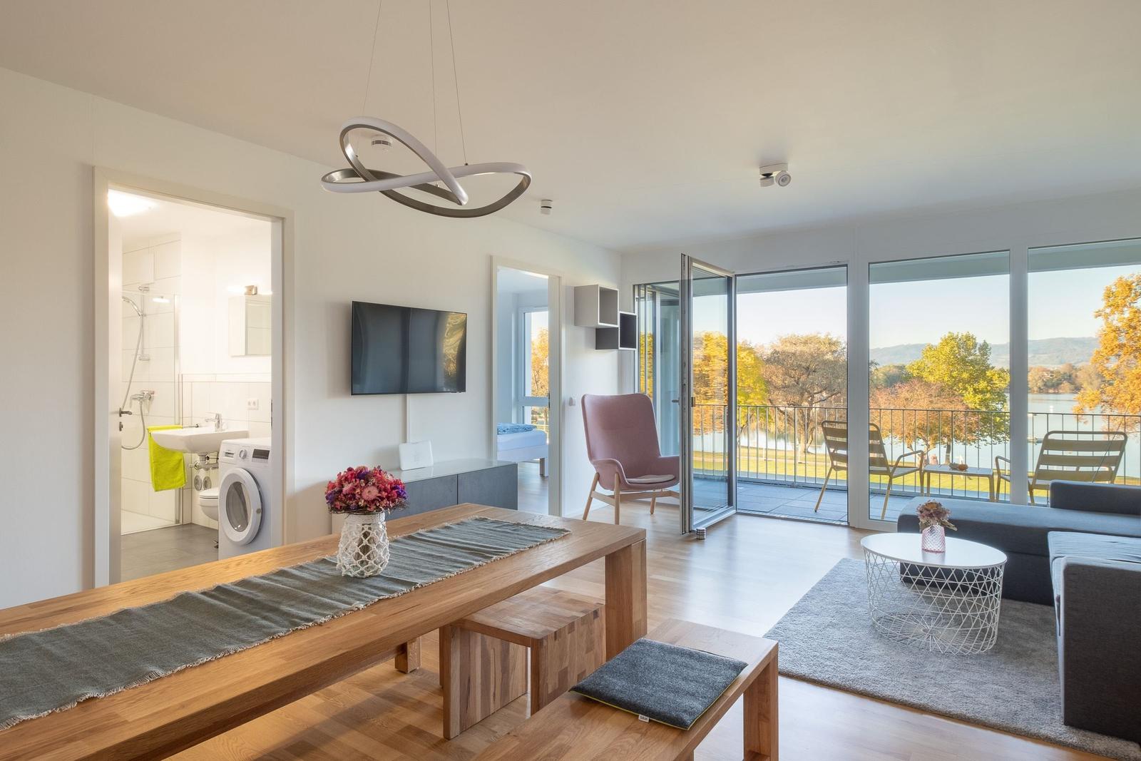Wohnraum mit Blick auf See, Esstisch, Sofa und Balkon mit Stühlen.