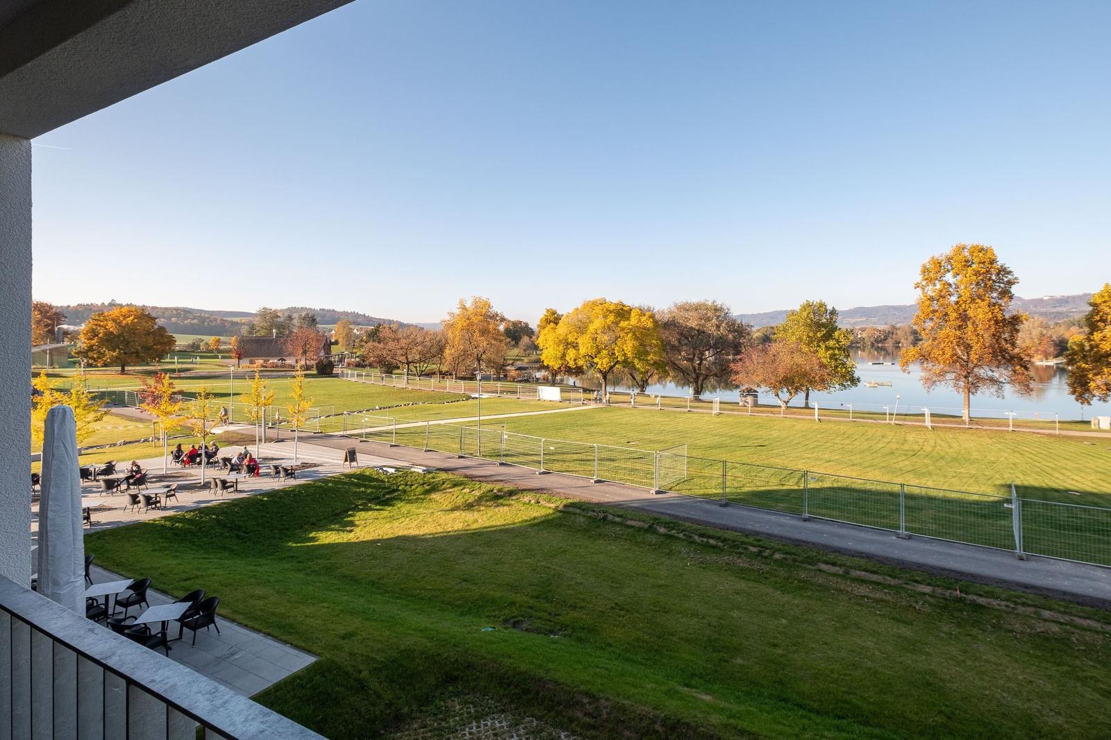 Blick von Balkon auf Grünfläche, See und Herbstbäume.