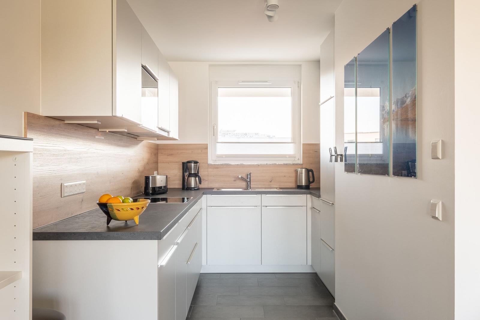 Moderne Küche mit weißen Schränken, dunkler Arbeitsplatte und Holzrückwand. Fenster und Küchenzeile sind sichtbar.