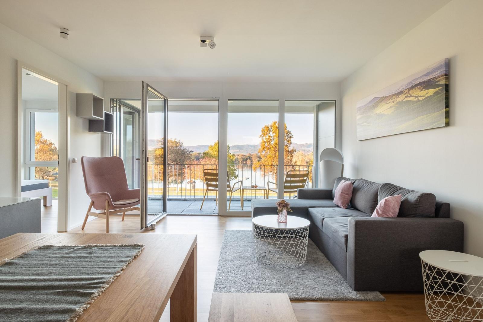 Wohnzimmer mit Sofa, Tisch und Balkonblick auf See und Hügel.