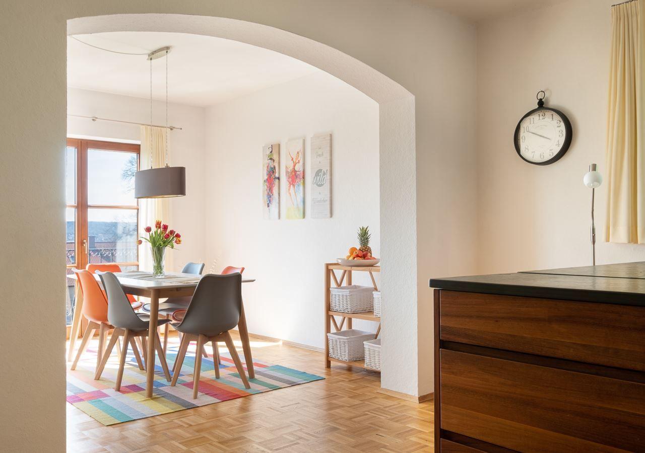 Essbereich mit Tisch, Stühlen und Fenster. Kücheninsel und Wanduhr sichtbar.