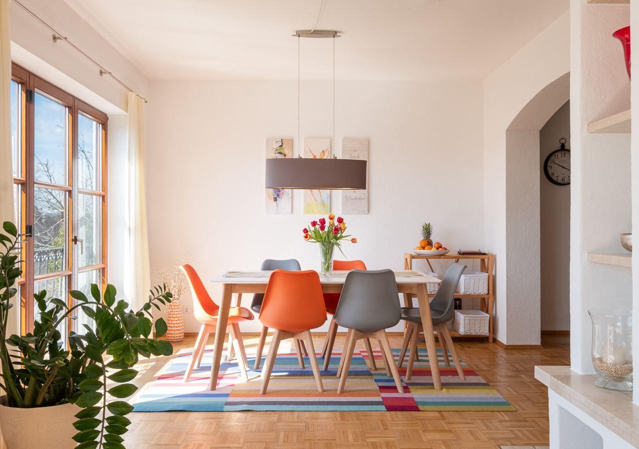 Esszimmer mit Holztisch, bunten Stühlen und buntem Teppich. Fenster, Pflanze und Wandbilder sichtbar.