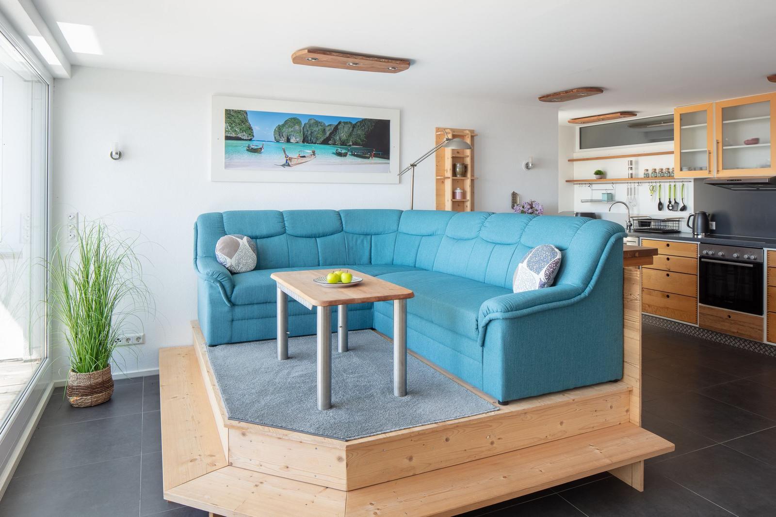Ein Wohnbereich mit blauem Sofa, Holzpodest und Küchenbereich im Hintergrund.