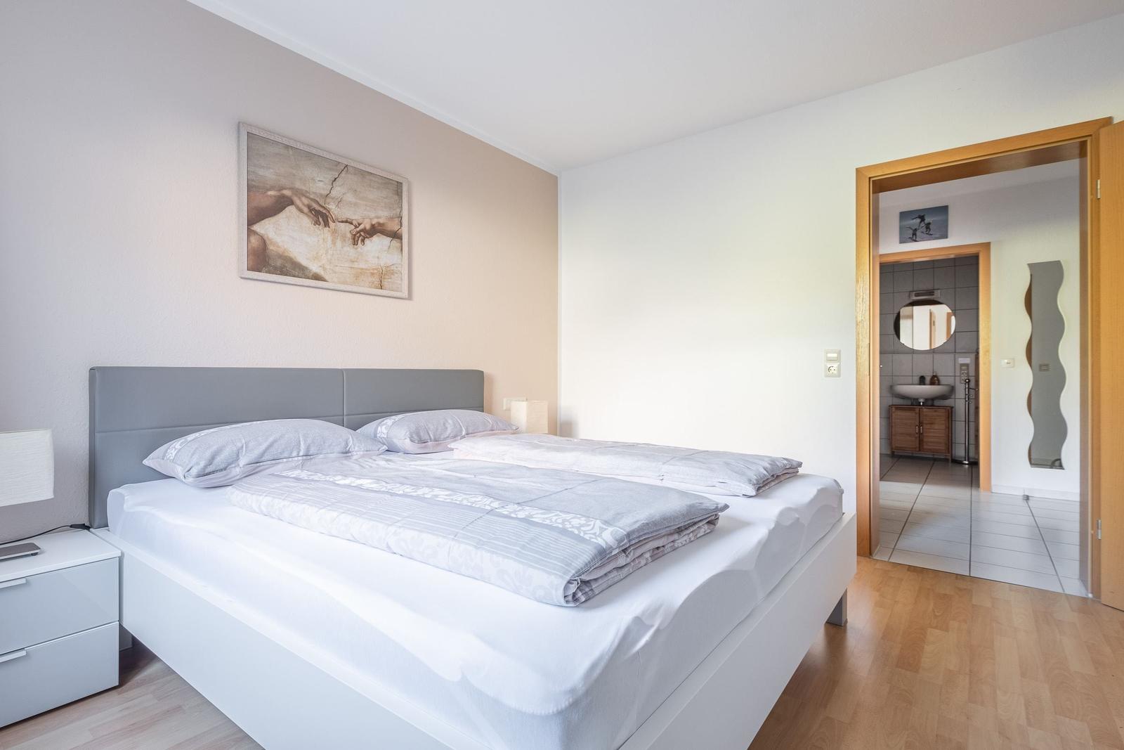 Schlafzimmer mit Doppelbett, Nachttisch und Bild an der Wand. Tür führt in die Badewanne.