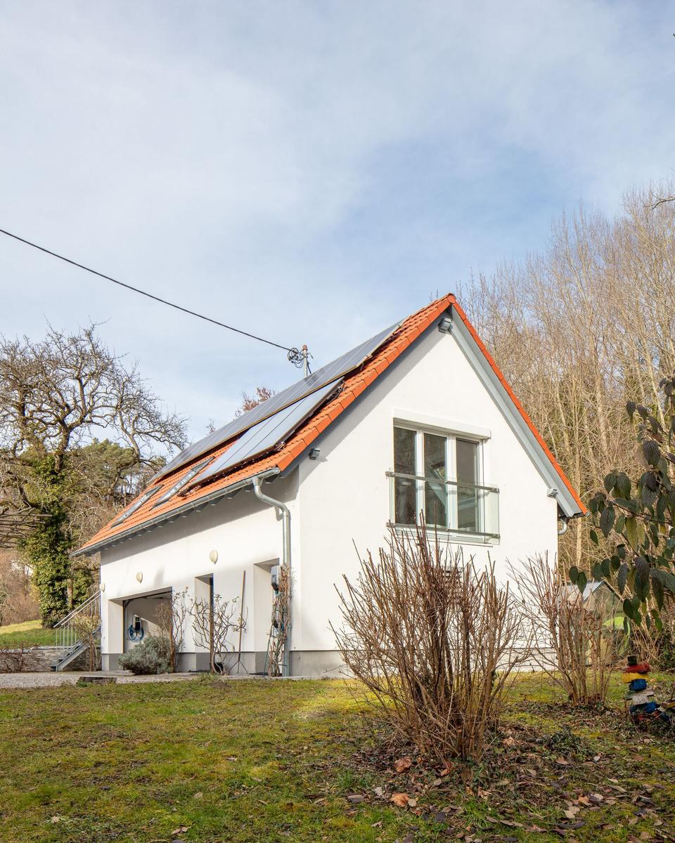 Ein weißes Haus mit rotem Dach und Solarpaneelen. Ein Balkon und ein Garagenbereich sind sichtbar.
