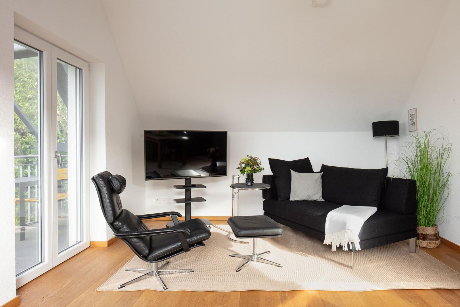 Ein Wohnzimmer mit schwarzem Sofa, Sessel und Fernseher. Holzboden und große Fenster.