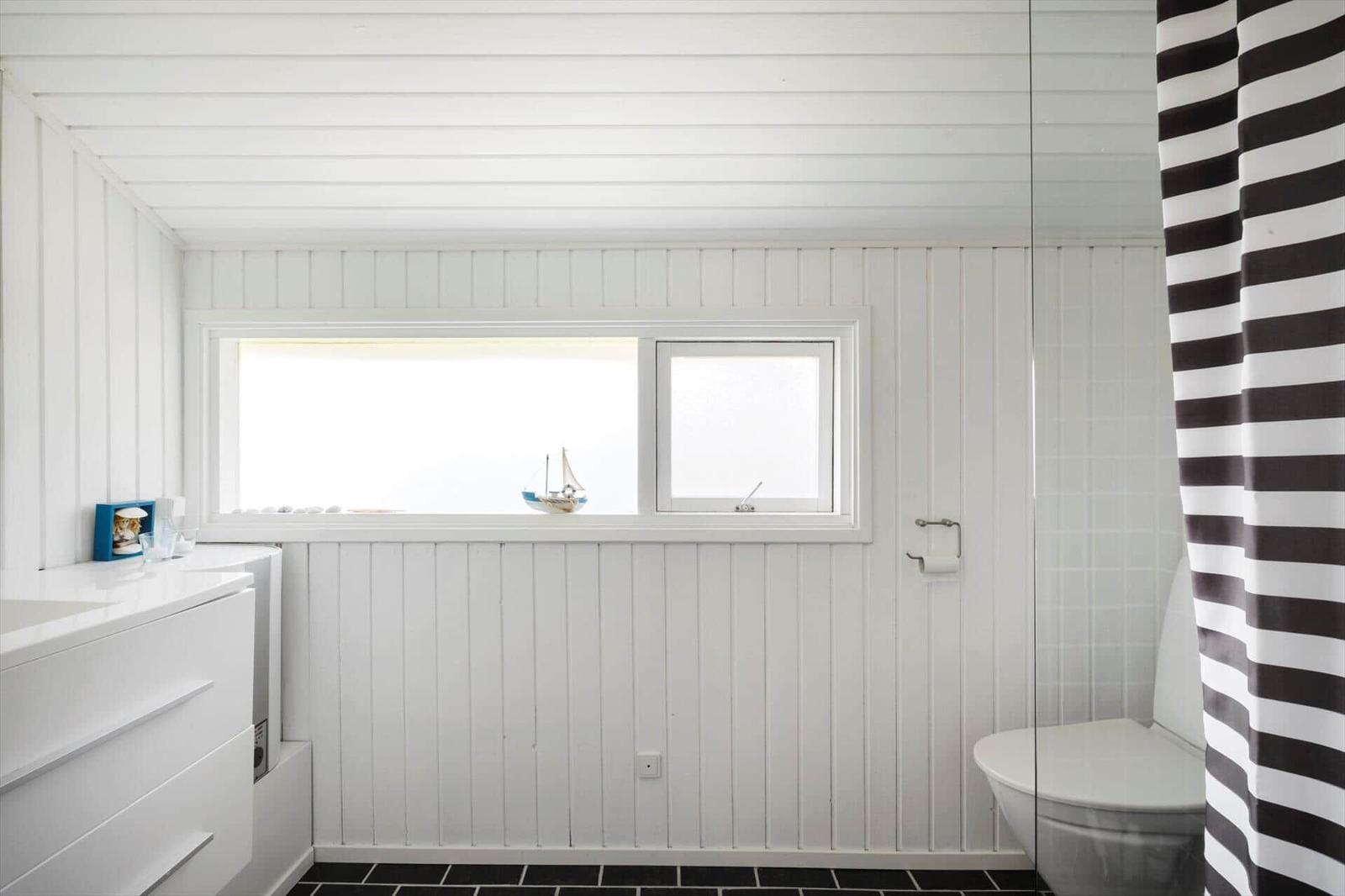 Badezimmer mit weißer Holzverkleidung, Fenster und schwarz-weißem Duschvorhang.