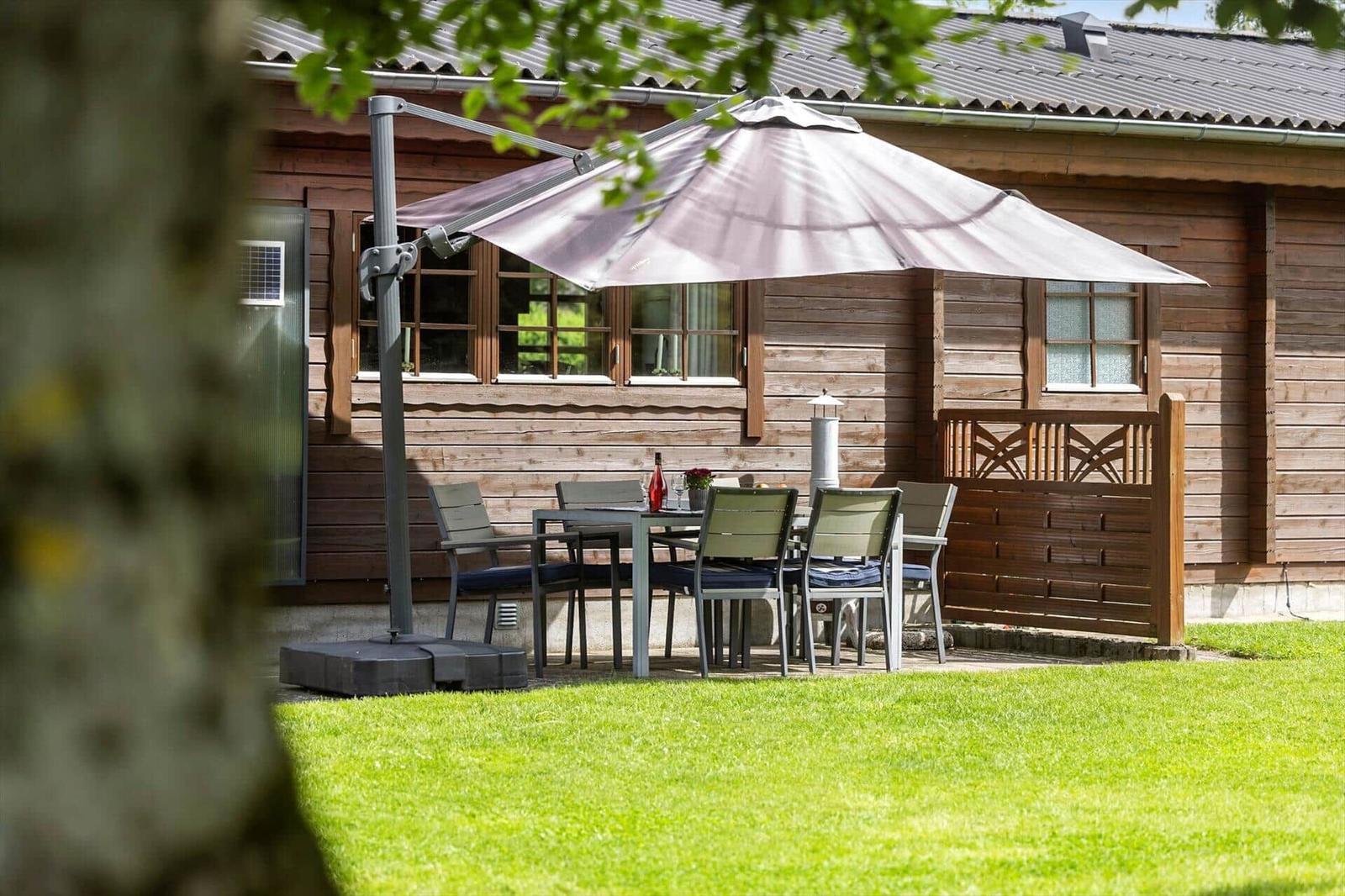 Terrasse mit Tisch, Stühlen und Sonnenschirm vor Holzhaus mit Grasfläche.