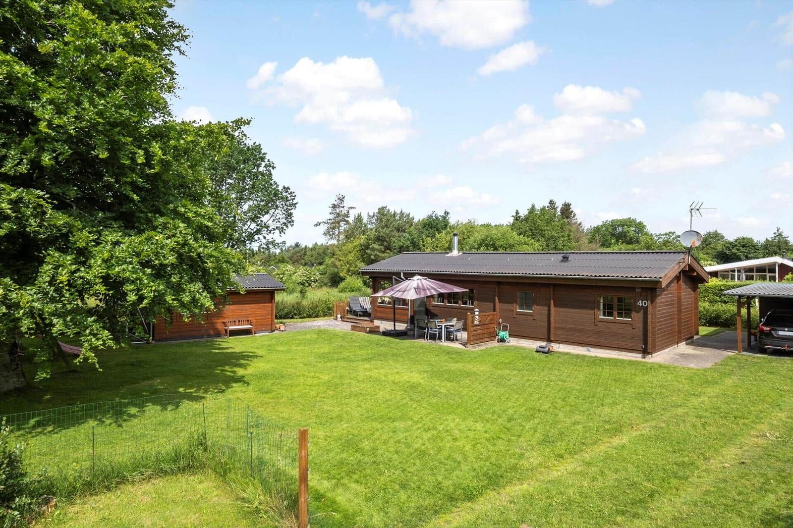Holzhaus mit Garten, Terrasse und Sonnenschutz. Links Nebengebäude, rechts Parkplatz.