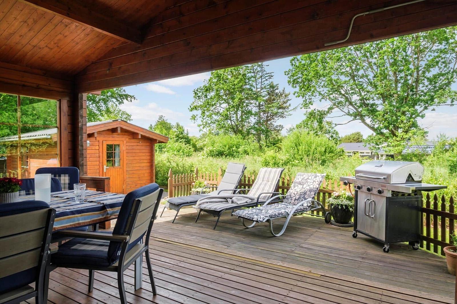 Terrasse mit Grill, Liegestühlen und Tisch unter Holzüberdachung. Blick auf grüne Landschaft.