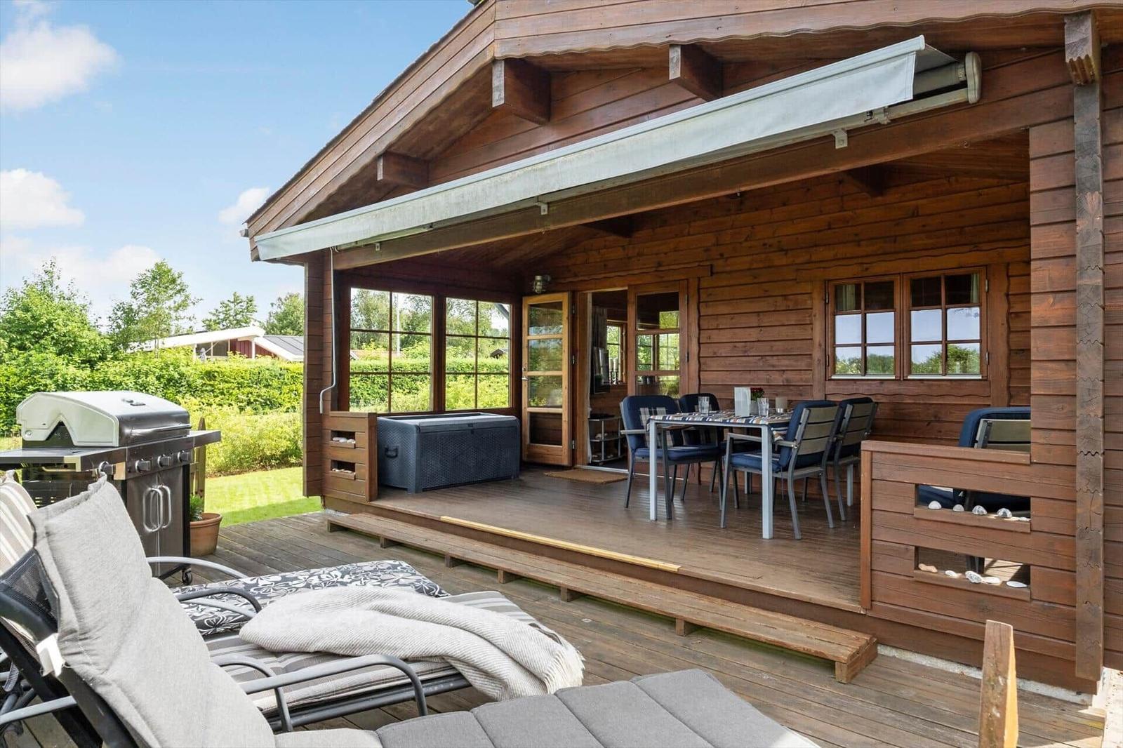 Terrasse mit Grill, Sitzgelegenheiten und Esstisch unter Markise. Holzhaus mit Fenstern und Türen.