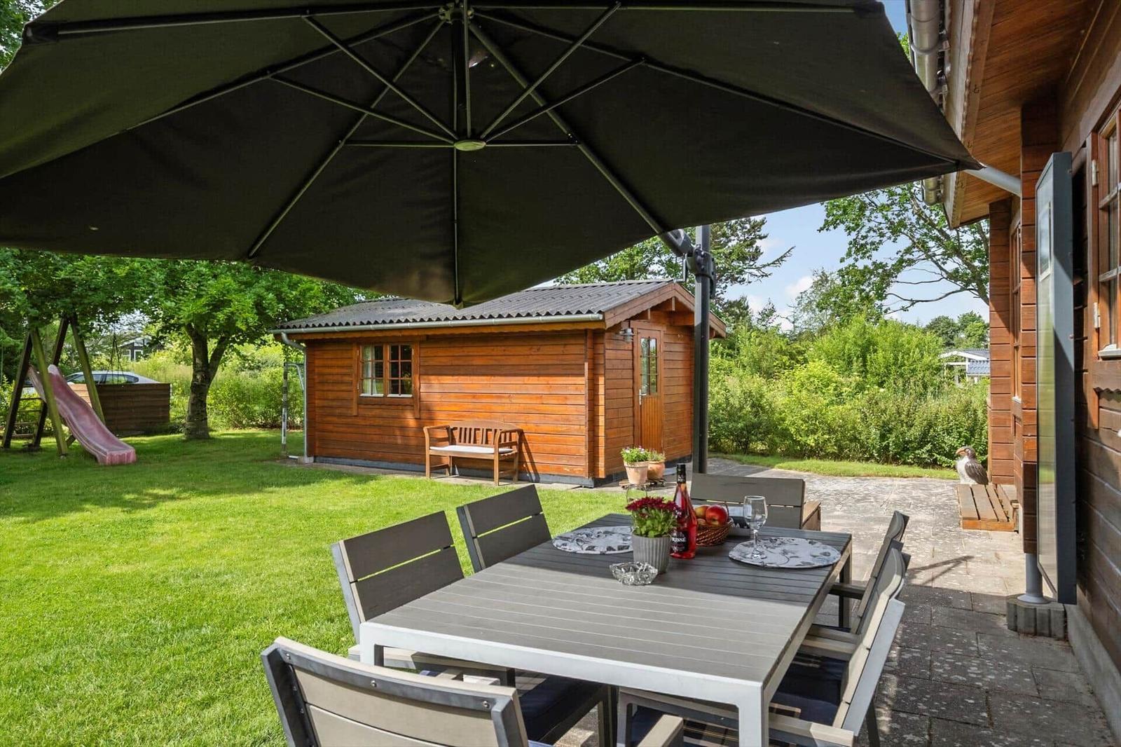 Terrasse mit Tisch, Stühlen und Sonnenschirm. Hinterhof mit Holzhaus und Spielplatz.