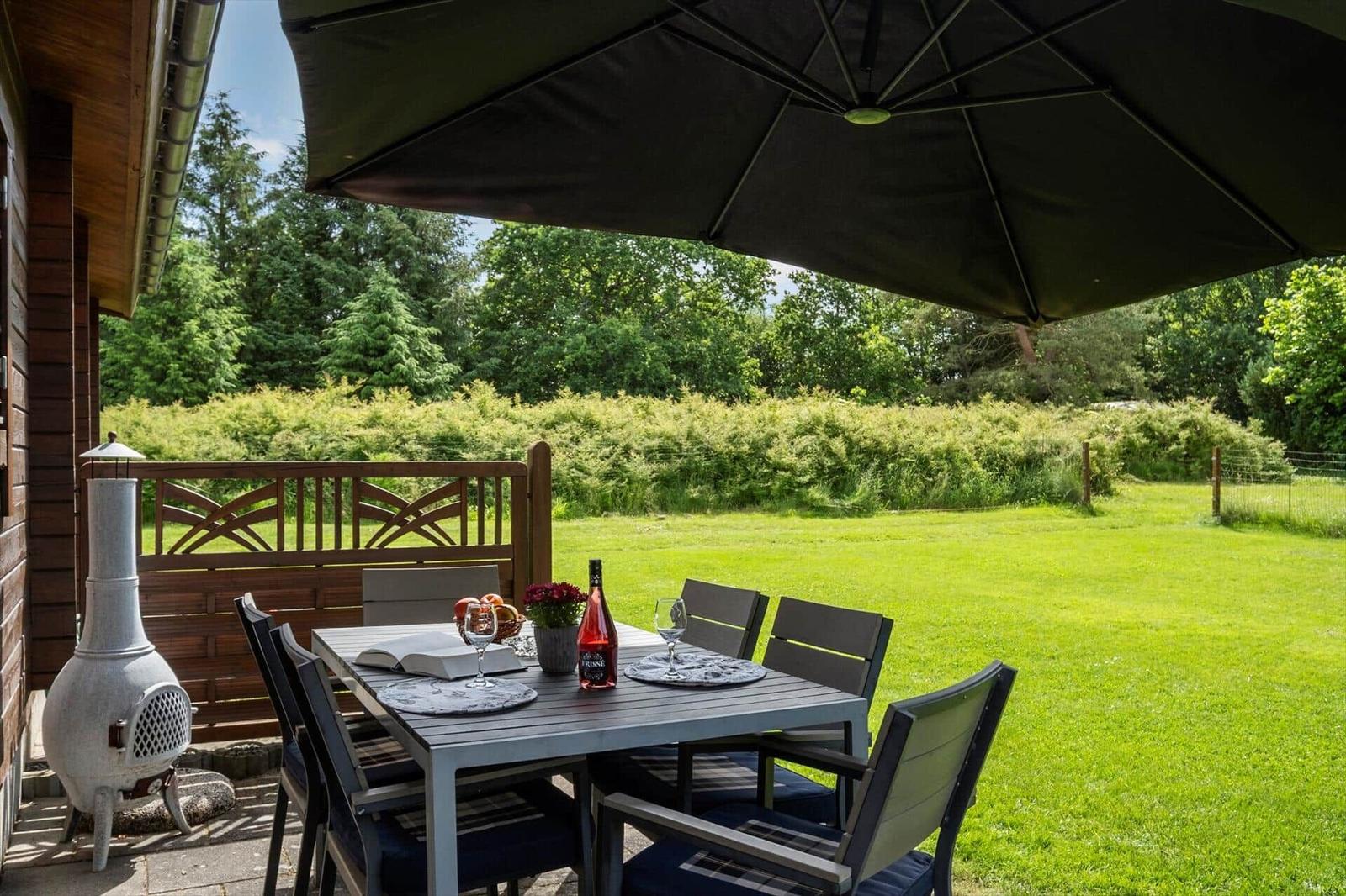 Terrasse mit Tisch, Stühlen und Sonnenschutz. Grünfläche und Hecke im Hintergrund.