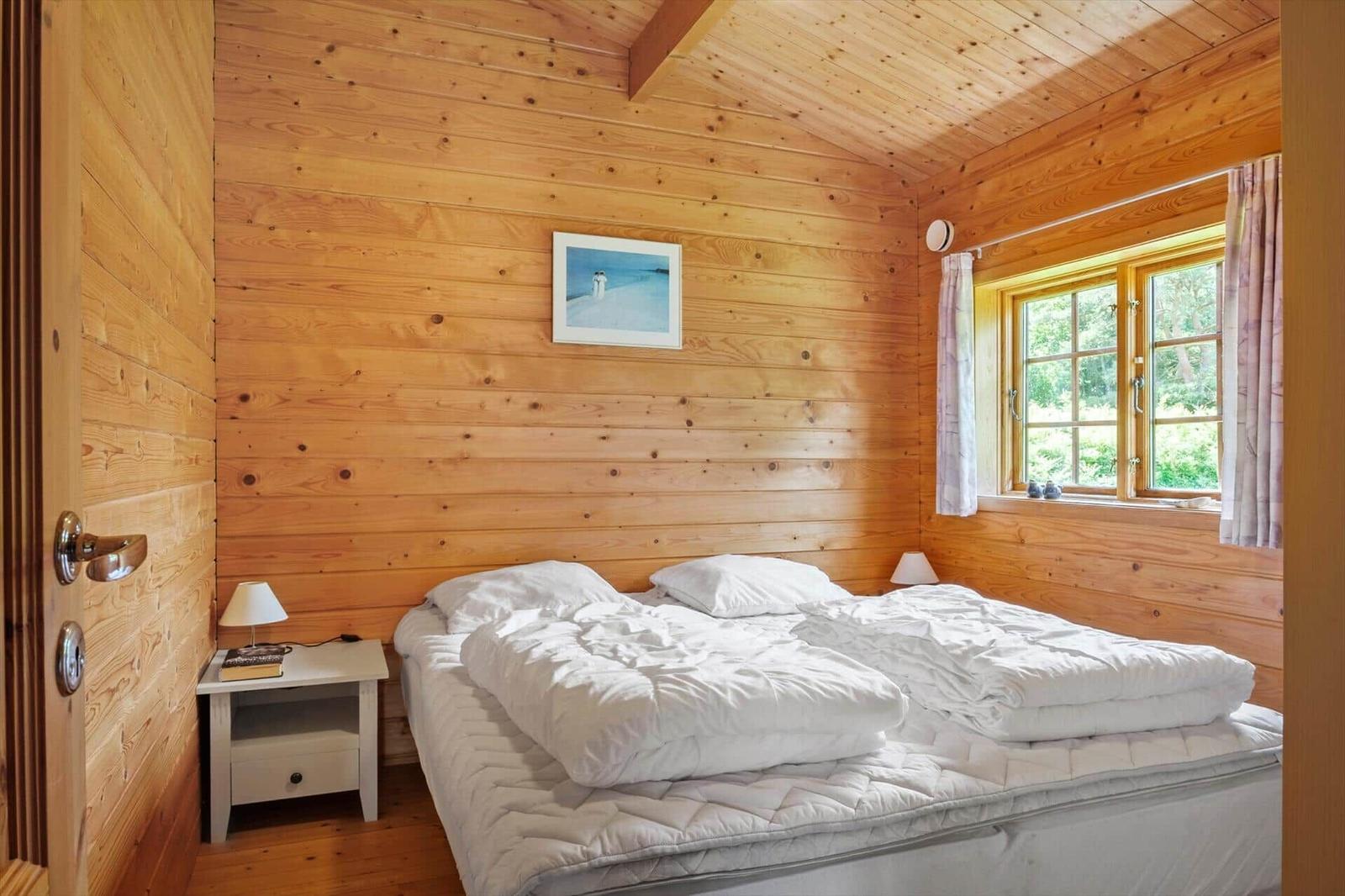 Schlafzimmer mit Holzwand, Bett und Fenster. Neben dem Bett steht ein Nachttisch.