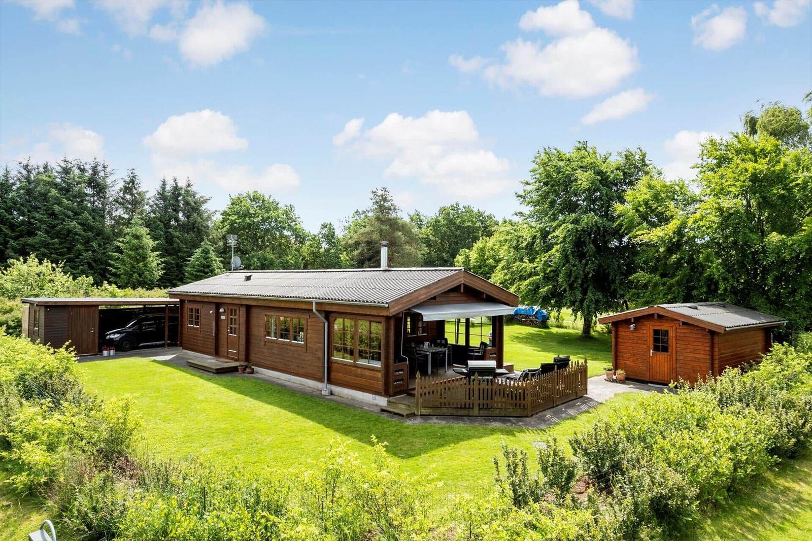 Holzhäuser mit Terrasse und Garten. Auto im Carport. Umgeben von Bäumen.