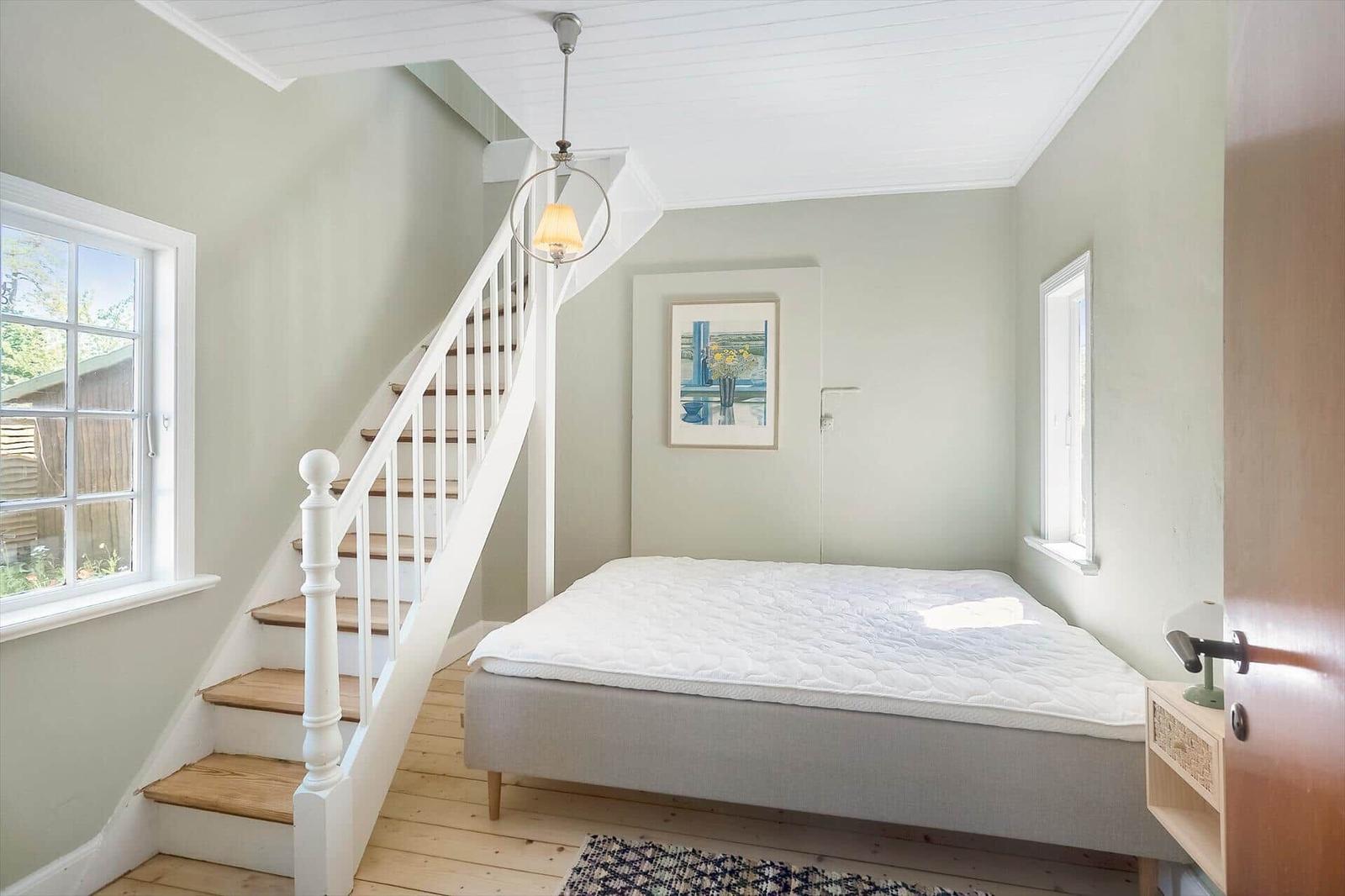 Schlafzimmer mit Bett, Treppe und Fenstern. Wandbild und Deckenleuchte sichtbar.