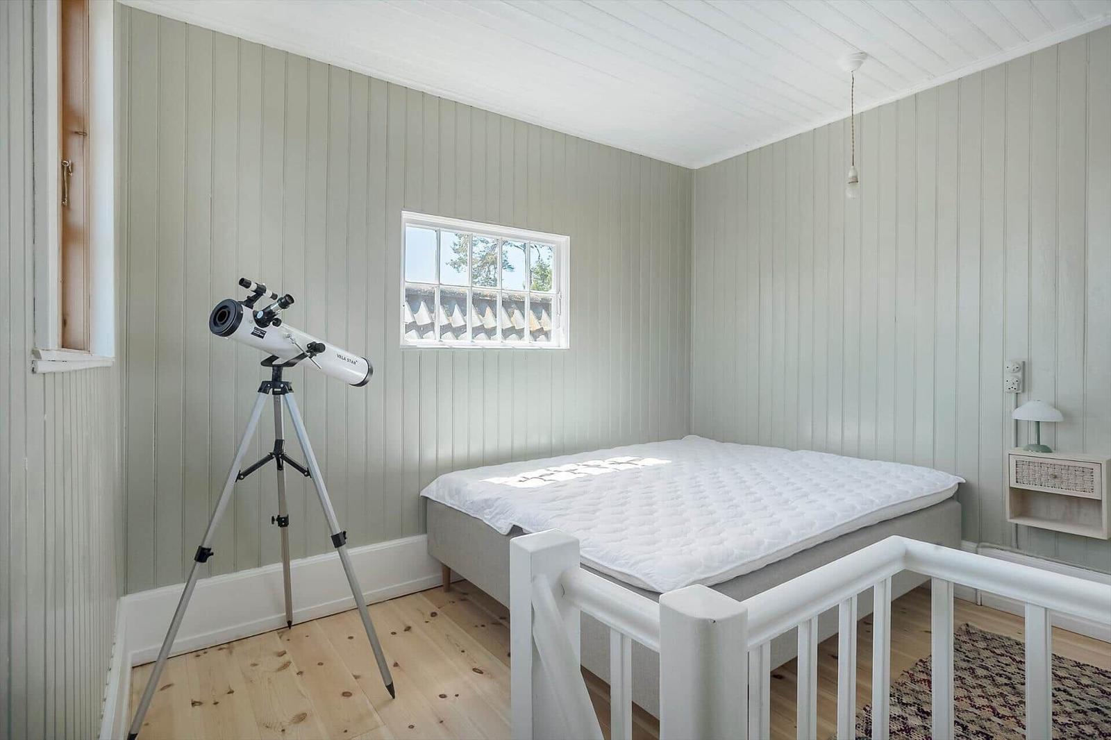 Schlafzimmer mit Bett, Teleskop und Fenster. Holzboden und Paneelen an den Wänden.