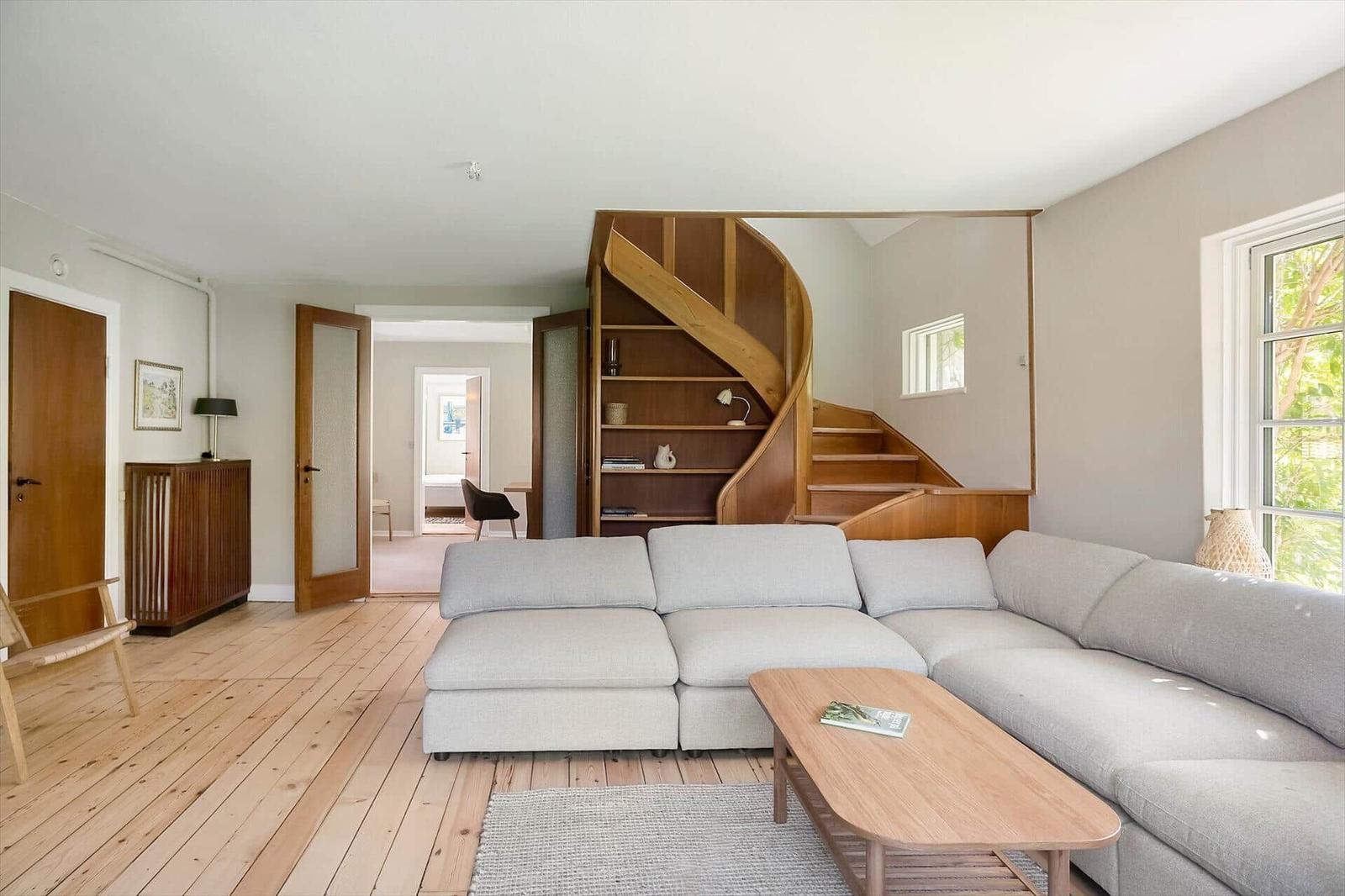 Wohnzimmer mit hellem Sofa, Holzfußboden und Wendeltreppe mit integrierten Regalen.