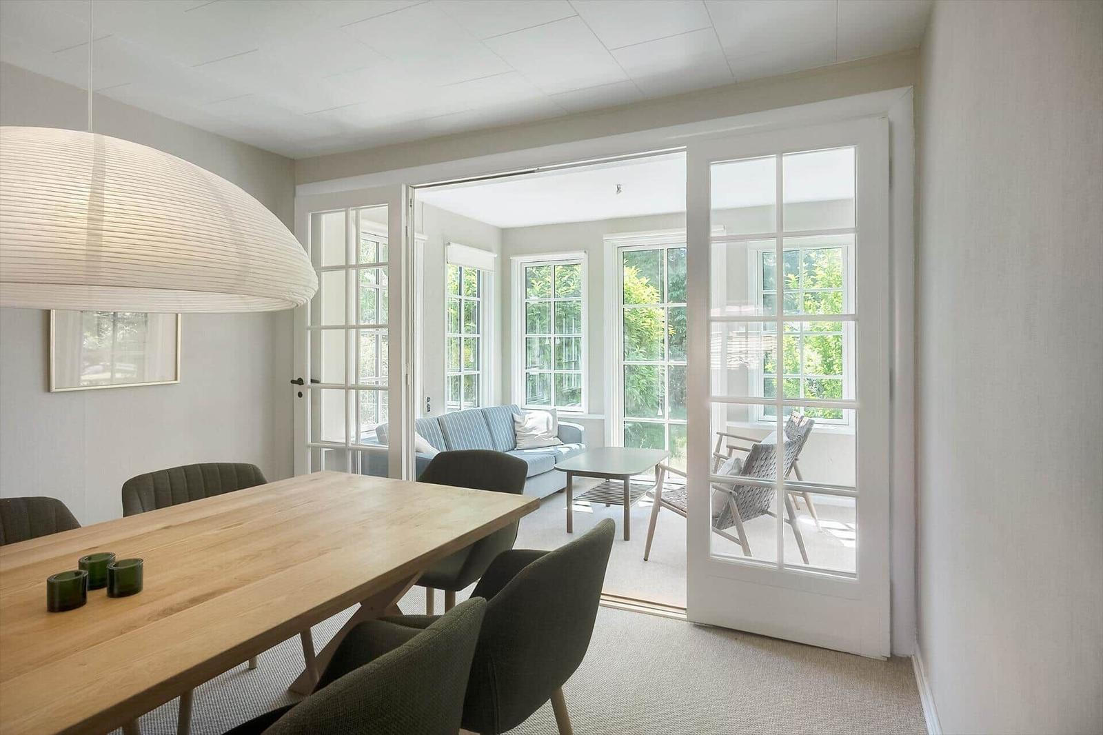 Esszimmer mit Tisch und Stühlen, durchgehendes Fenster zum Wohnbereich mit Sofa und Gartenblick.