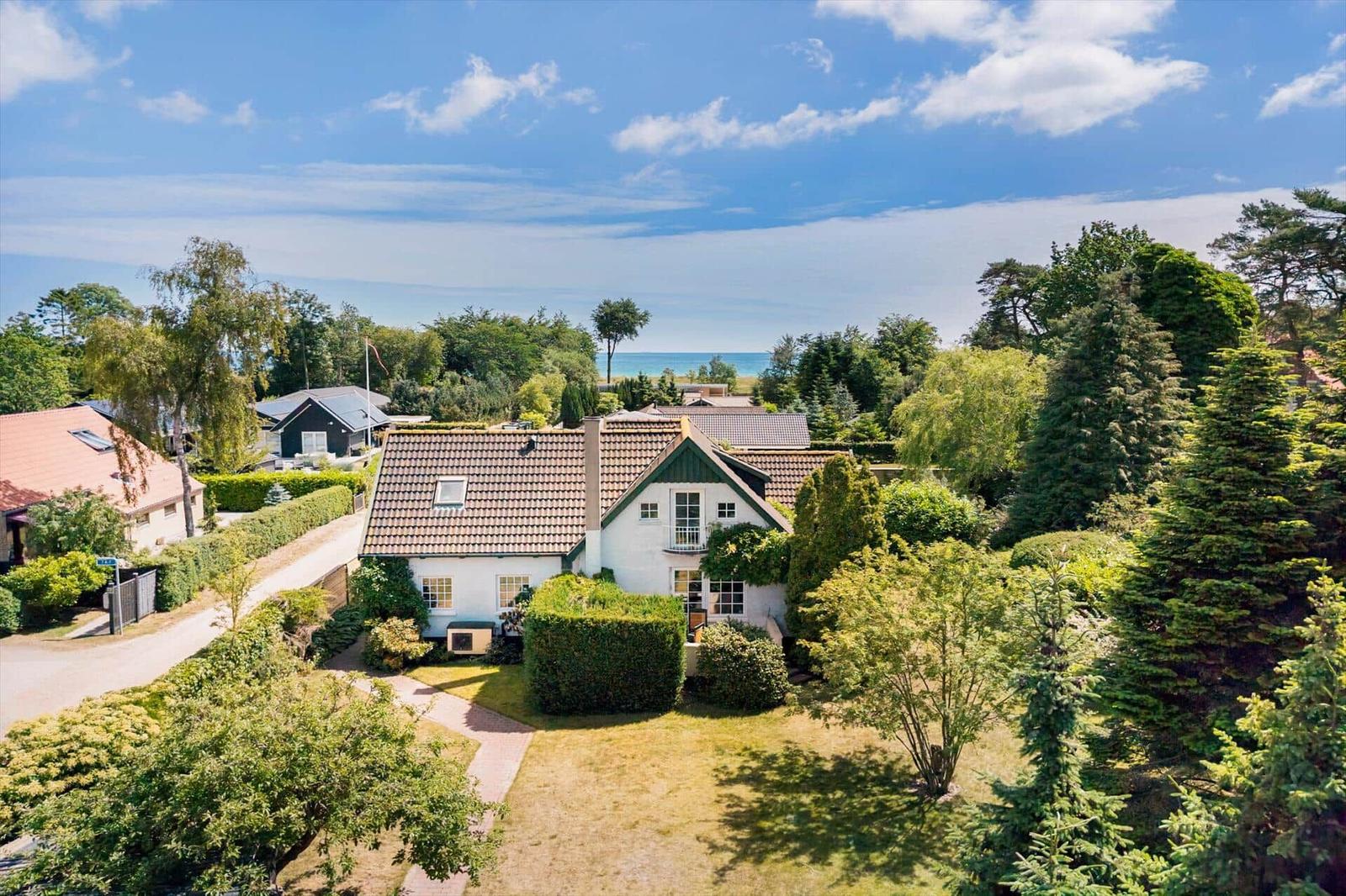 Ein weißes Haus mit braunem Dach, umgeben von Grünflächen und Bäumen, mit Blick auf das Meer.