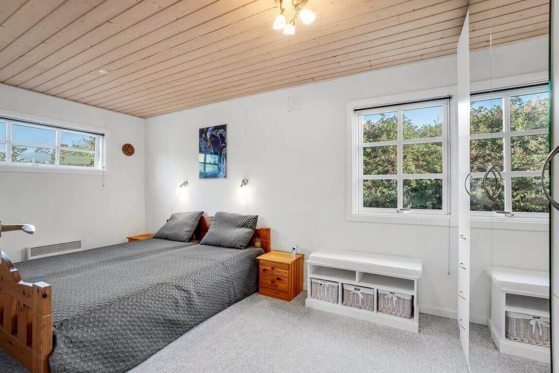 Schlafzimmer mit Holzdecke, Bett, Fenstern und Schrank. Weiße Wände und Holzmöbel.
