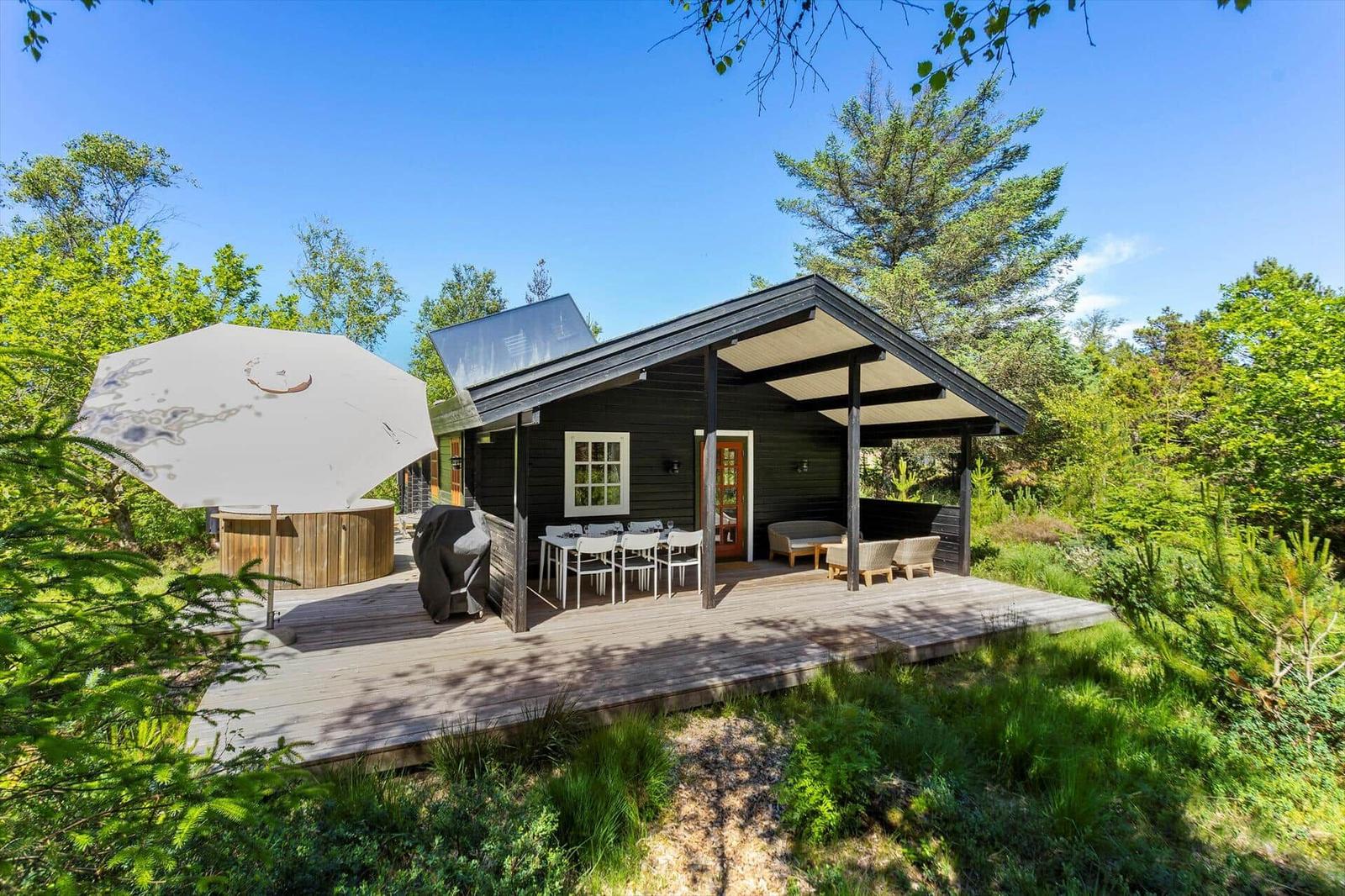 Holzhaus mit Terrasse, Gartenmöbeln und Sonnenschirm. Umgeben von Bäumen und Gras.
