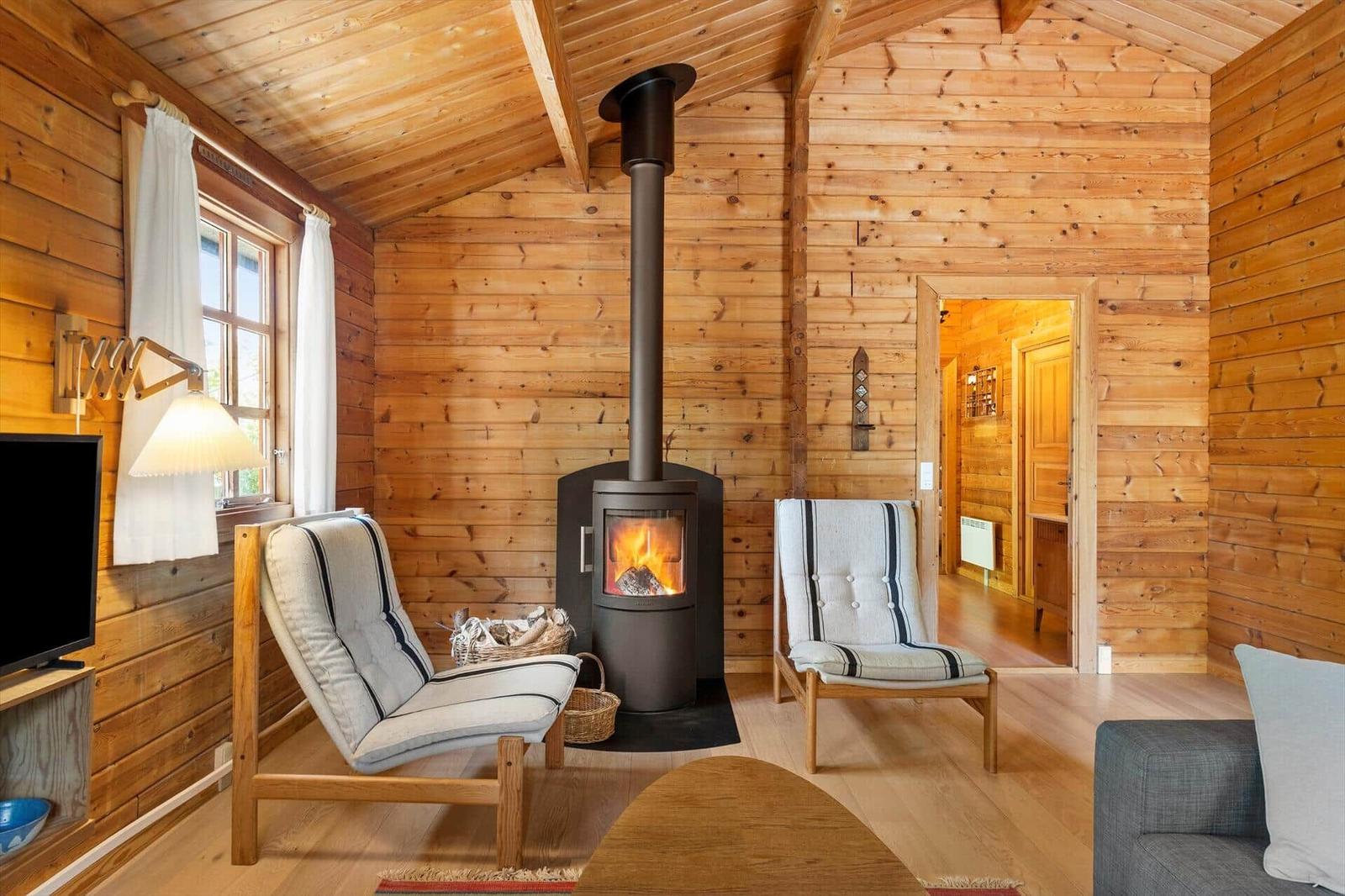Holzhaus mit Kamin, zwei Sesseln und Fenster. Tür führt in angrenzenden Raum.