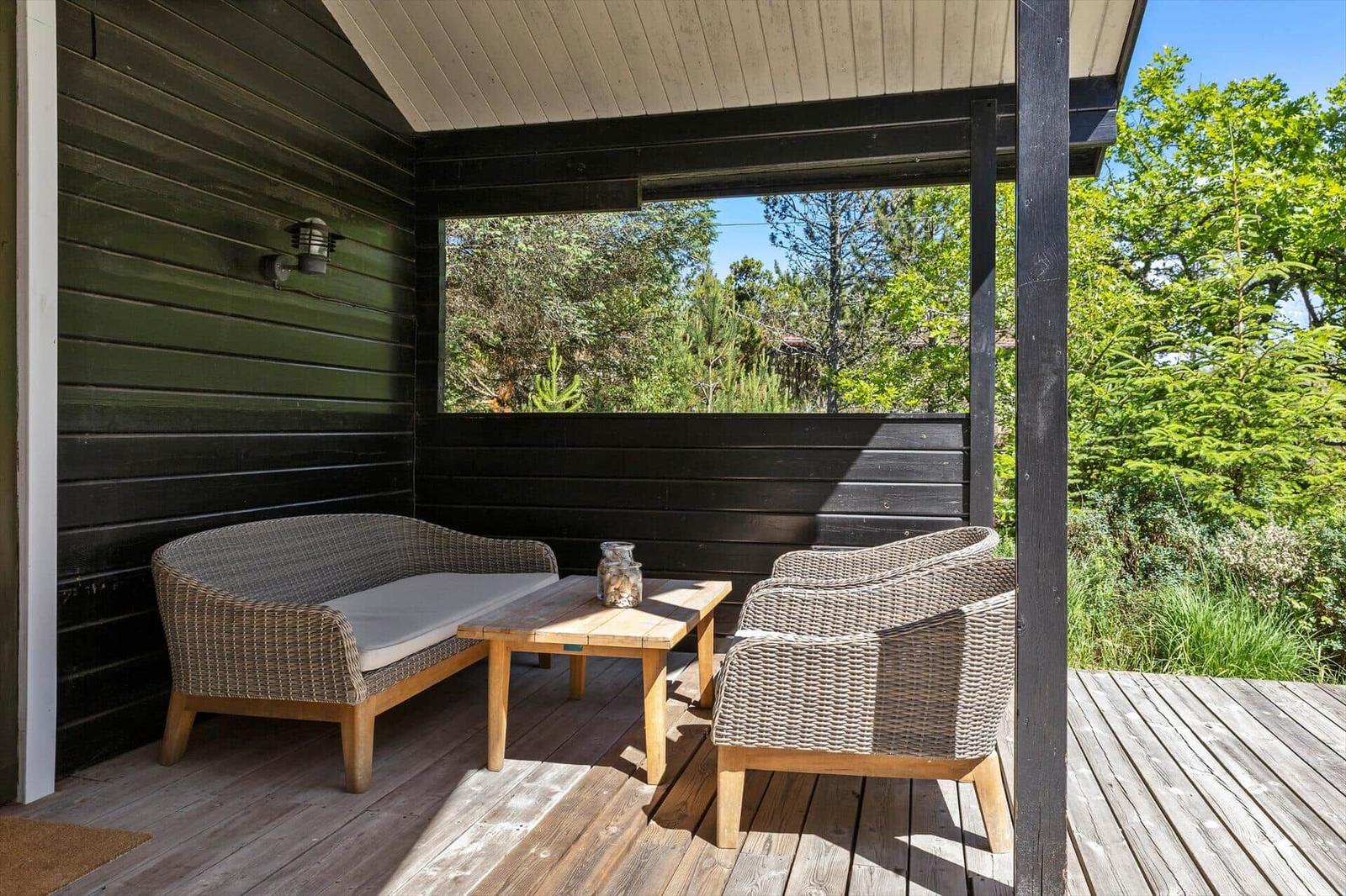 Terrasse mit Rattanmöbeln und Holztisch unter einem Dach mit Blick auf grüne Bäume.