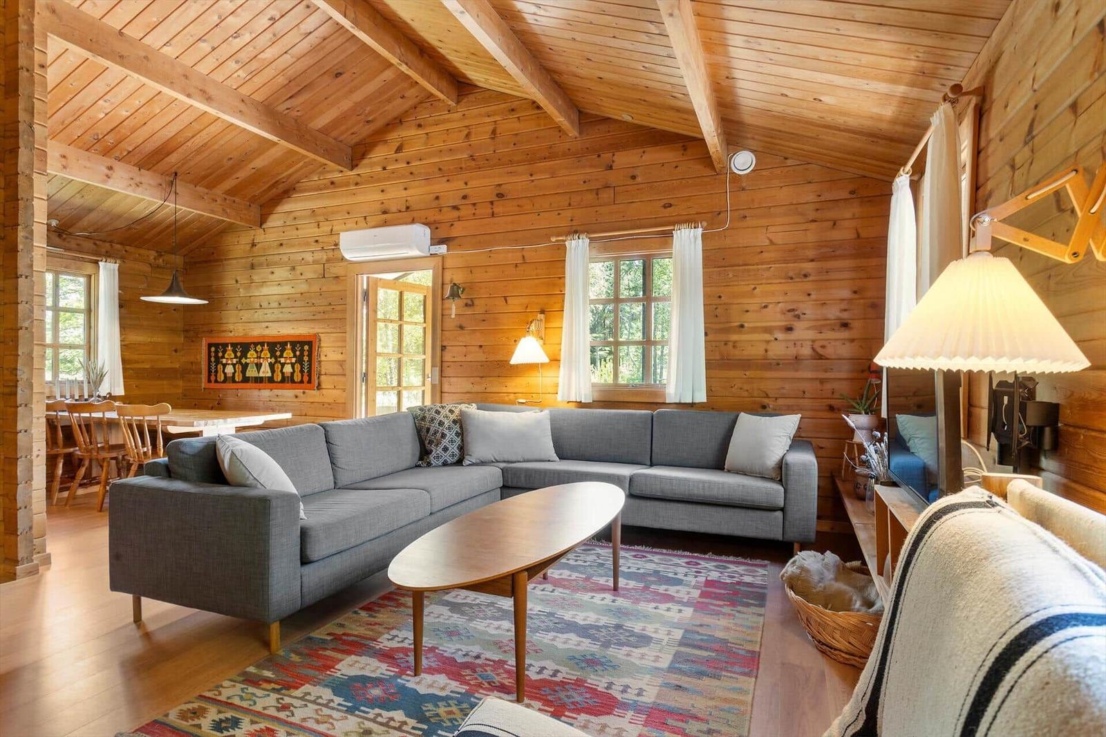 Wohnzimmer mit Holzwand, grauem Sofa und Holztisch auf gemustertem Teppich.