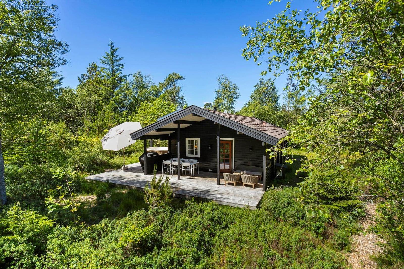 Holzhütte mit Terrasse, Gartenmöbeln und Sonnenschirm in grüner Umgebung.