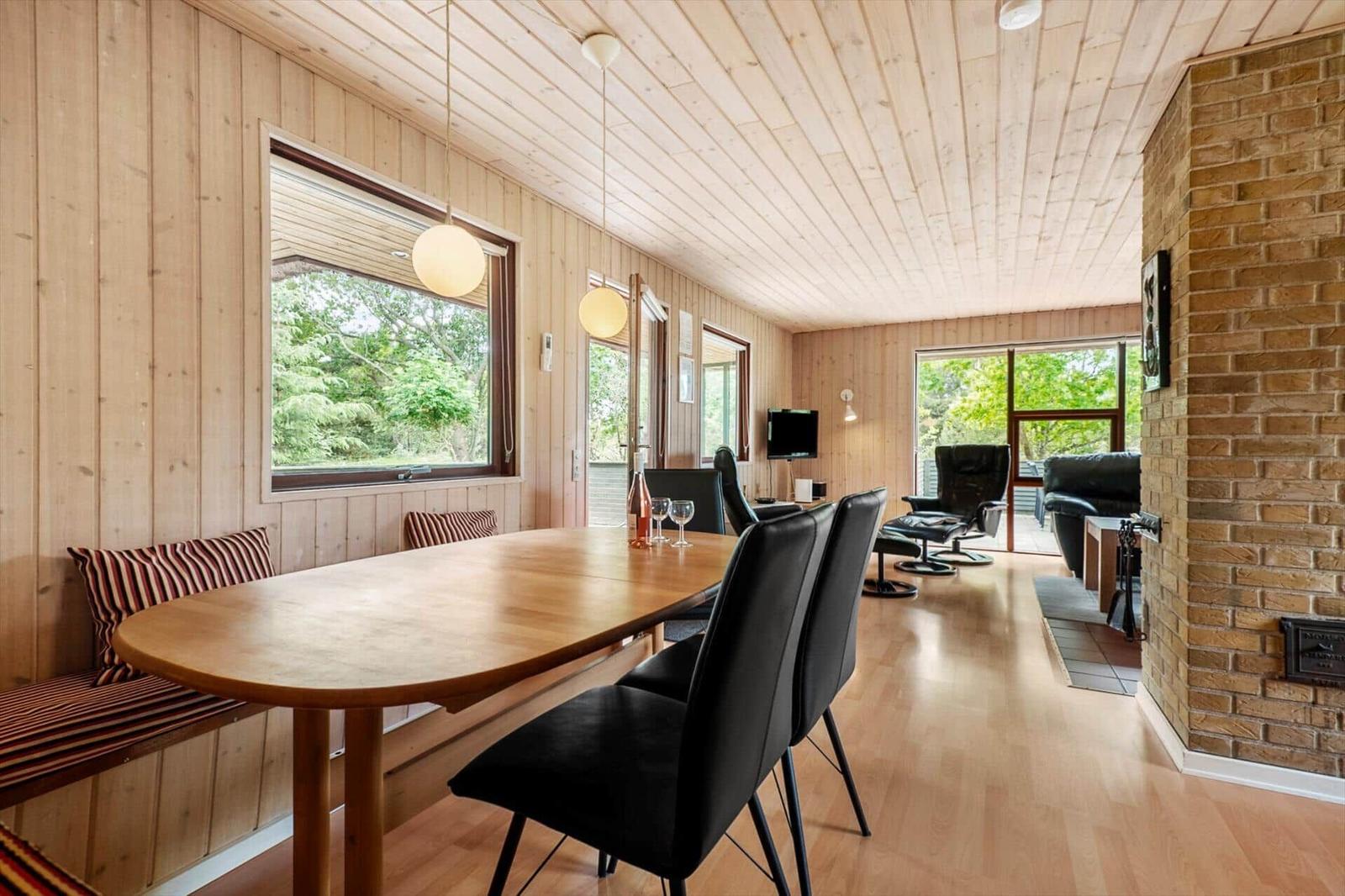 Essbereich mit Holztisch und schwarzen Stühlen. Im Hintergrund Wohnbereich mit Sitzmöbeln und Kamin.