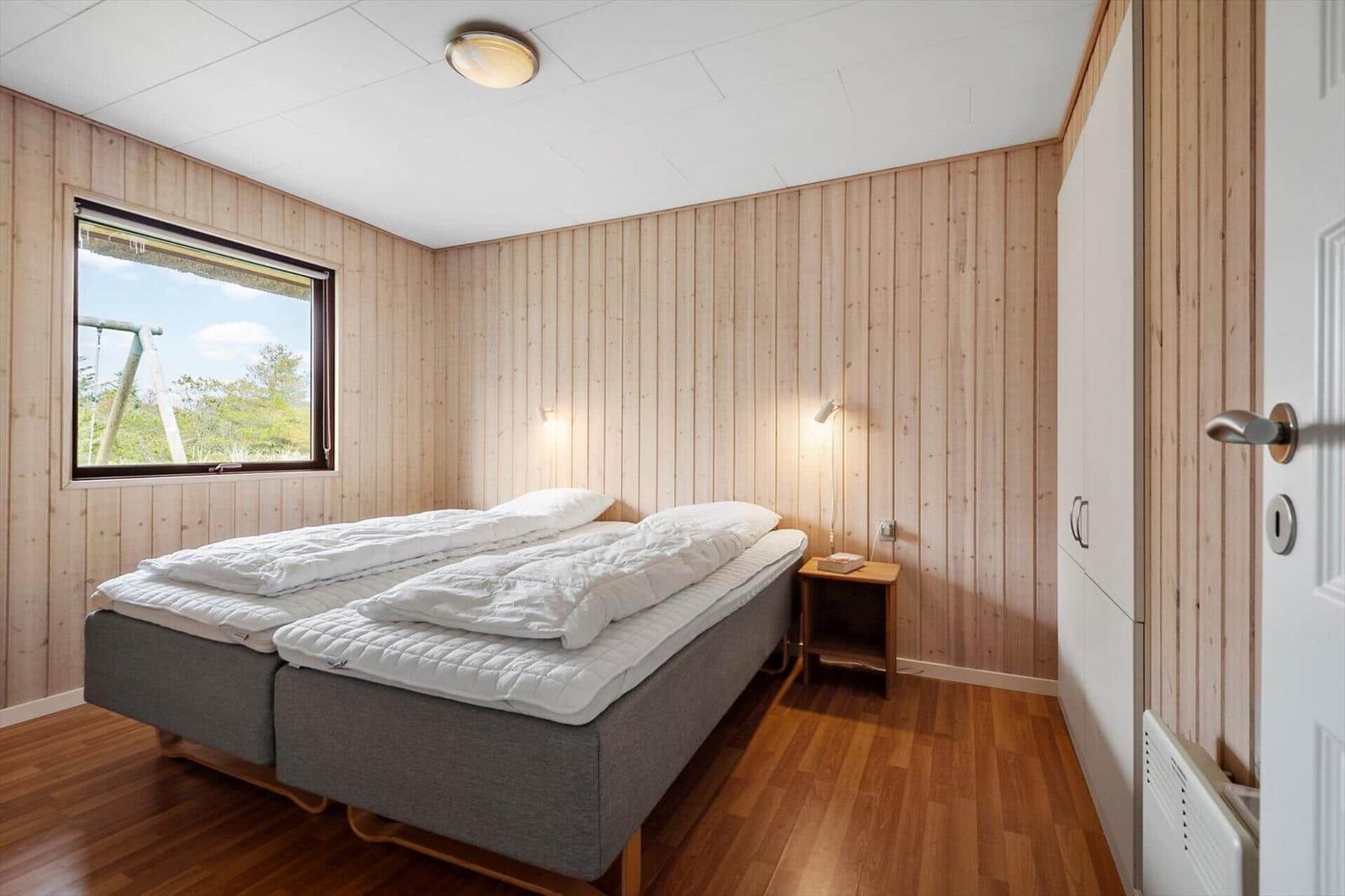 Schlafzimmer mit Doppelbett, Holzwand und Fensterblick auf Außenbereich.