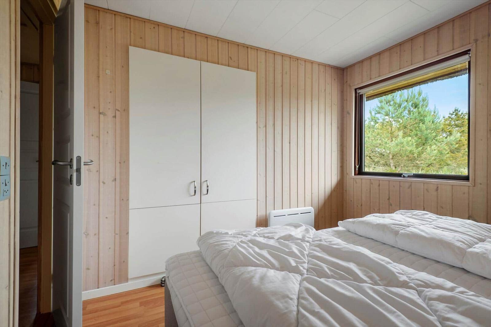 Schlafzimmer mit Bett, Kleiderschrank und Fenster mit Blick auf Wald.