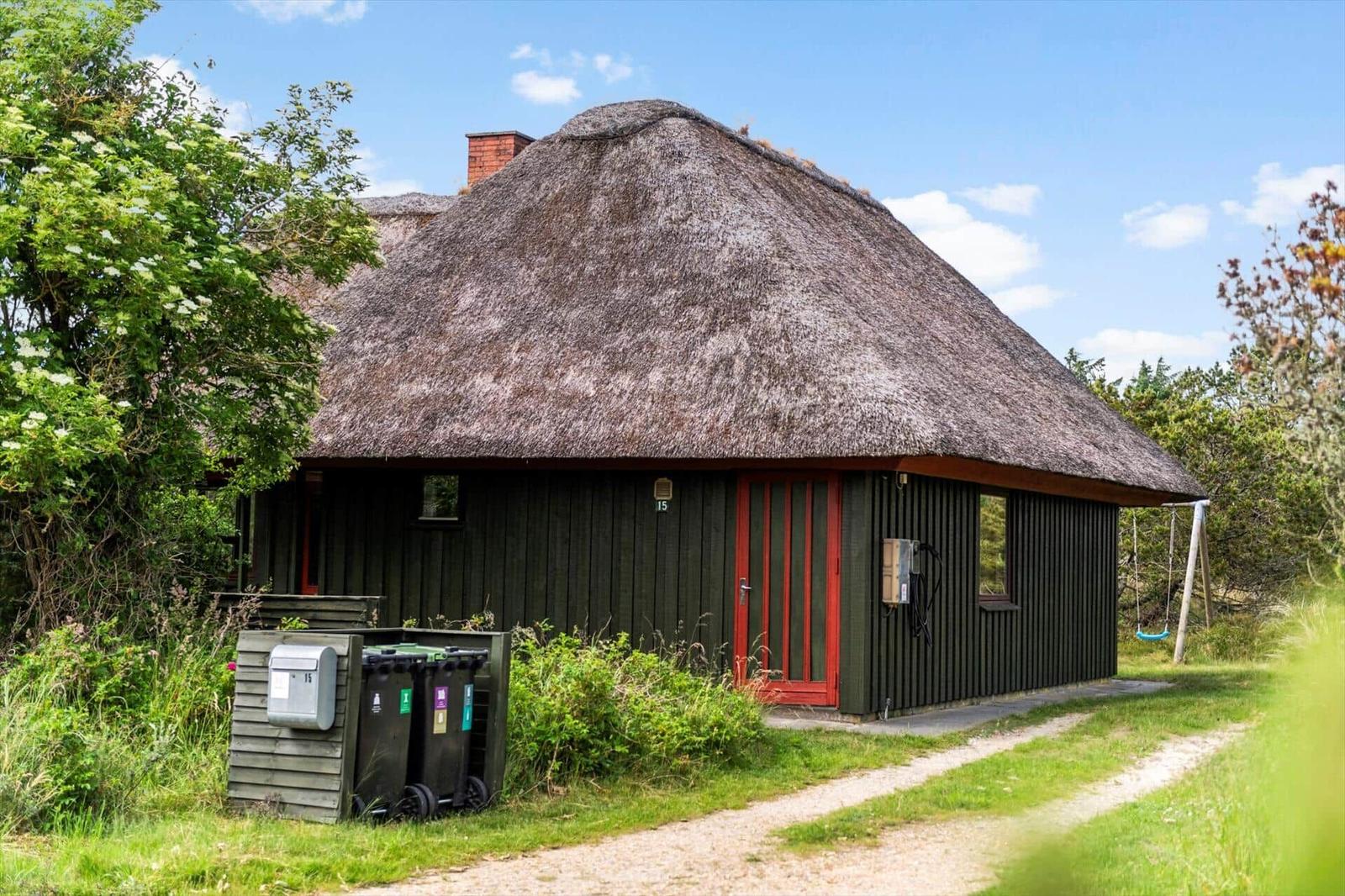 Haus mit strohgedecktem Dach, roter Tür und Umgebung mit Gras und Bäumen.