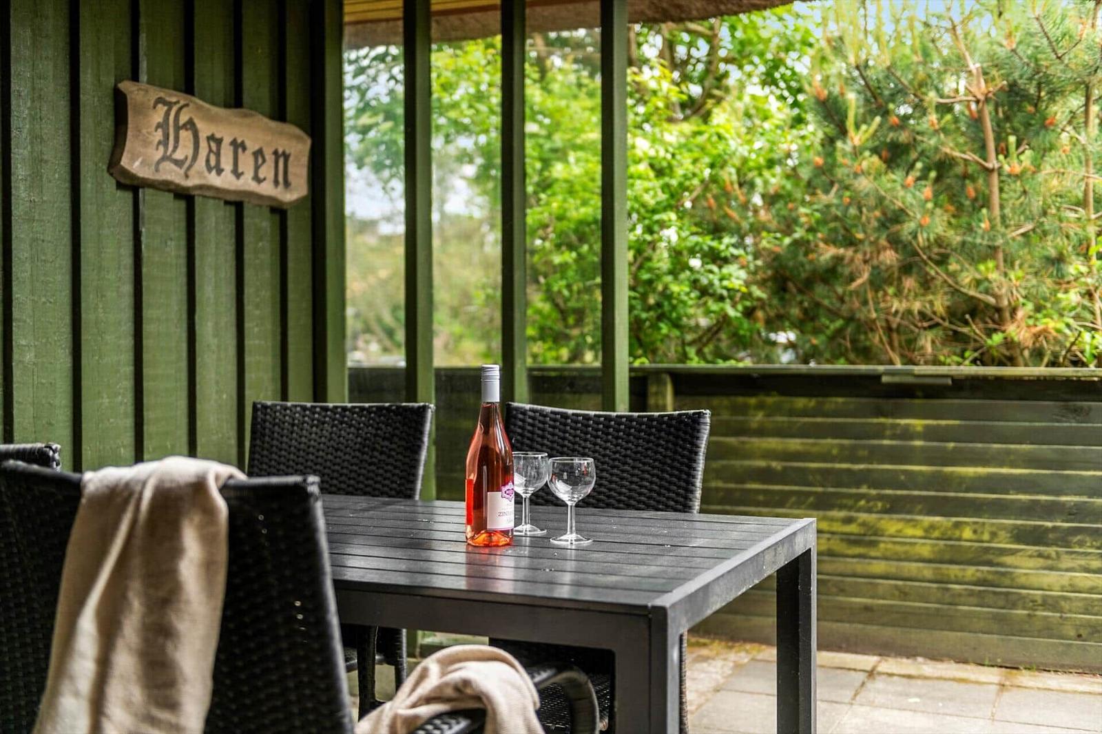Terrasse mit Tisch, Stühlen und einer Flasche Wein. Hintergrund: grüne Vegetation.