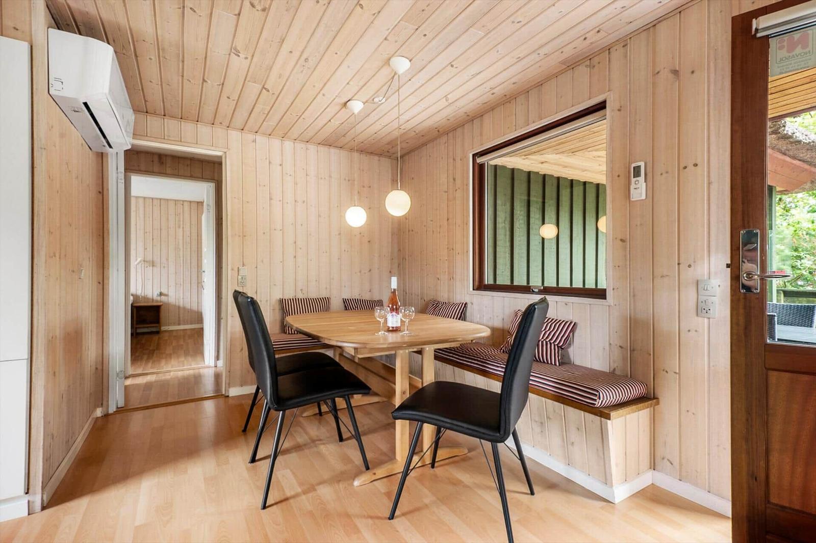 Esszimmer mit Holzwand, Tisch, Sitzbank und zwei Stühlen. Fenster und Tür sichtbar.