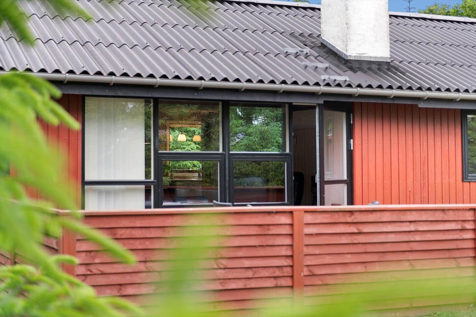 Rotes Holzhaus mit großem Fenster und Dach aus Wellblech. Im Inneren sind Möbel und Pflanzen sichtbar.