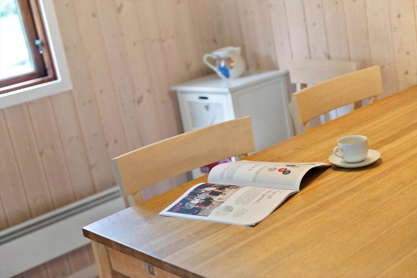 Holztisch mit Stuhl, Tasse und Zeitschrift. Holzwände und Fenster im Hintergrund.