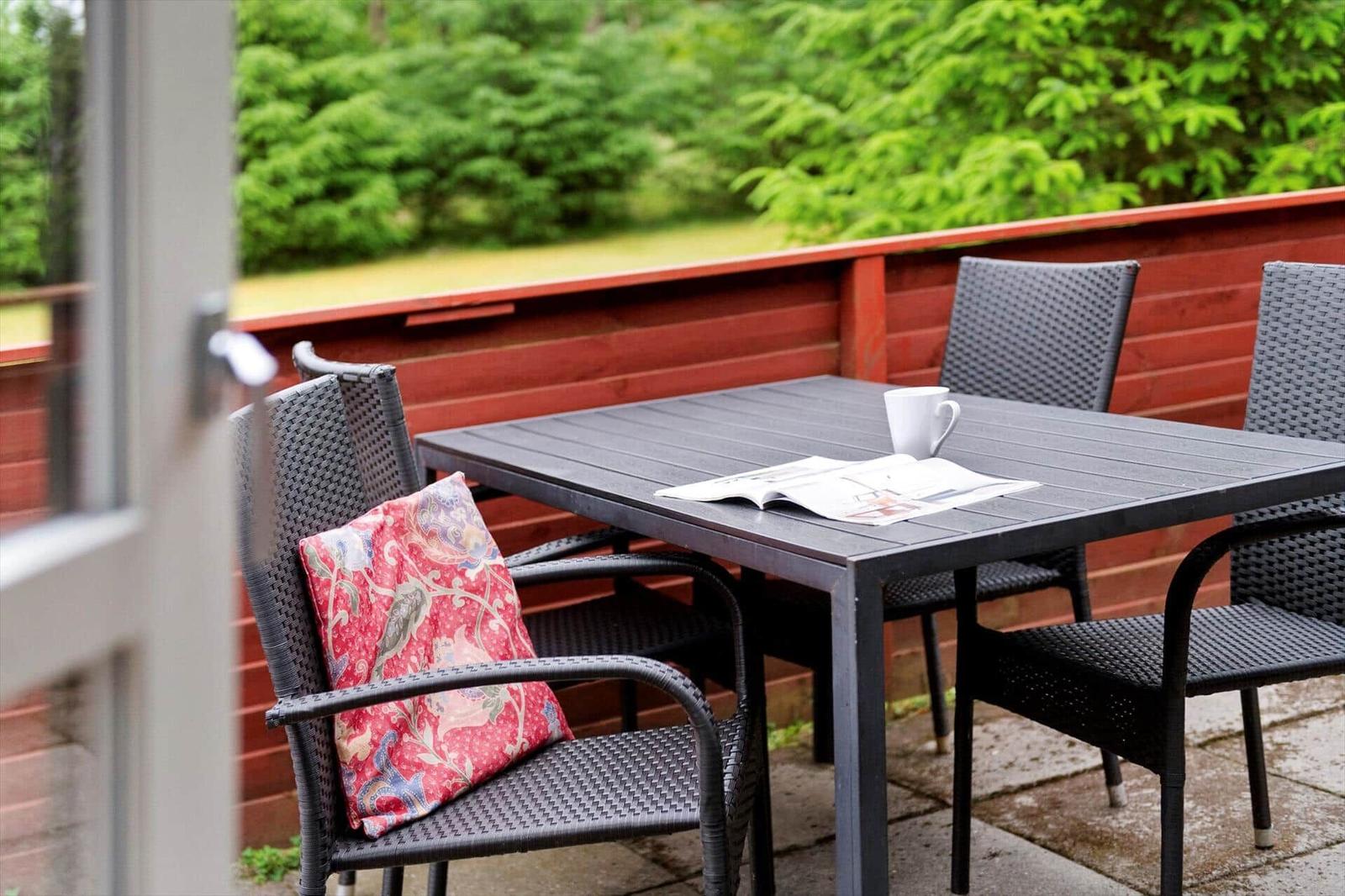 Terrasse mit Tisch, Stühlen und Kissen. Hintergrund: grüne Bäume und rote Holzablage.