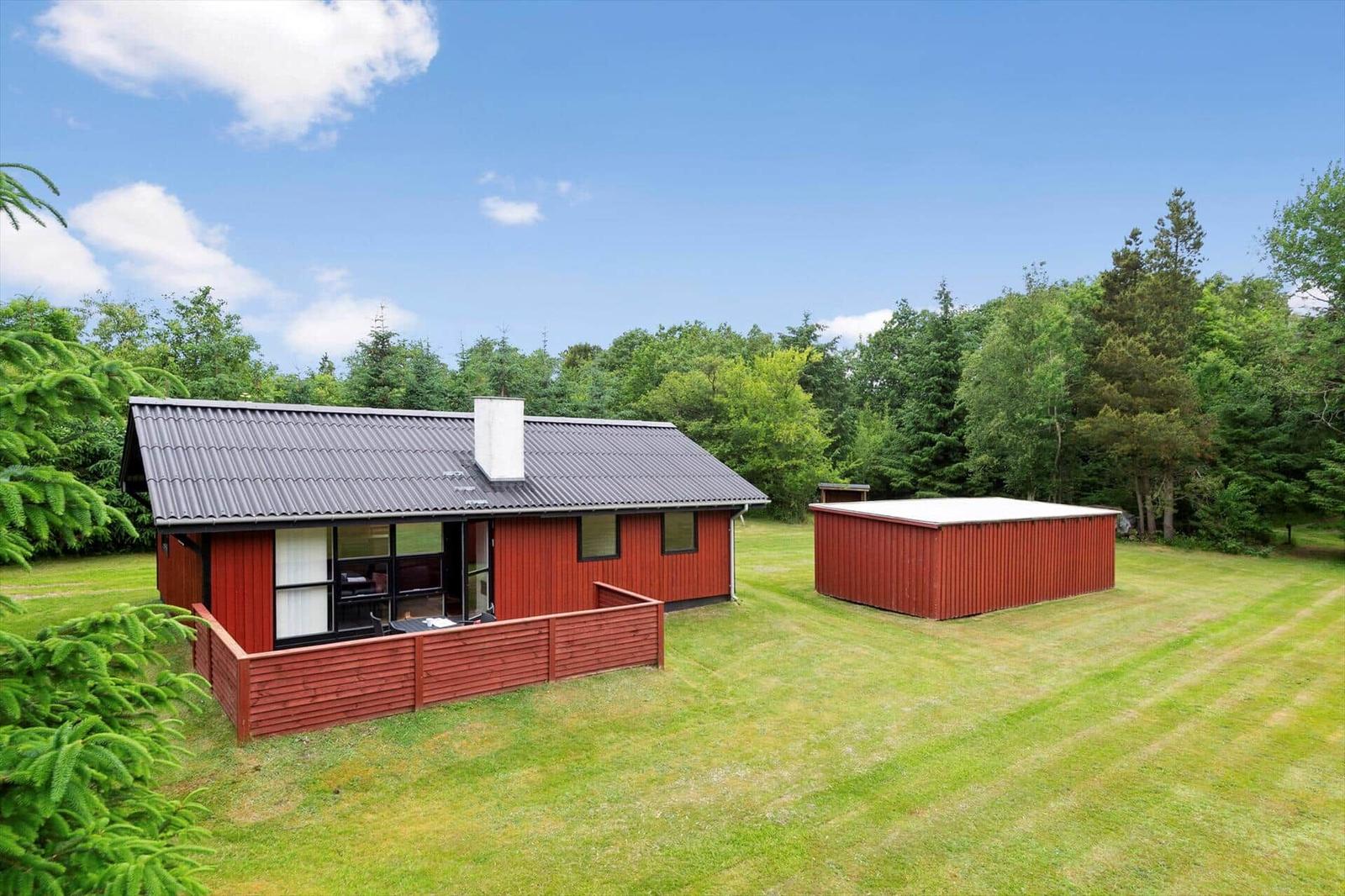 Rotes Holzhaus mit Grillplatz und großer grüner Wiese. Umgeben von Bäumen.
