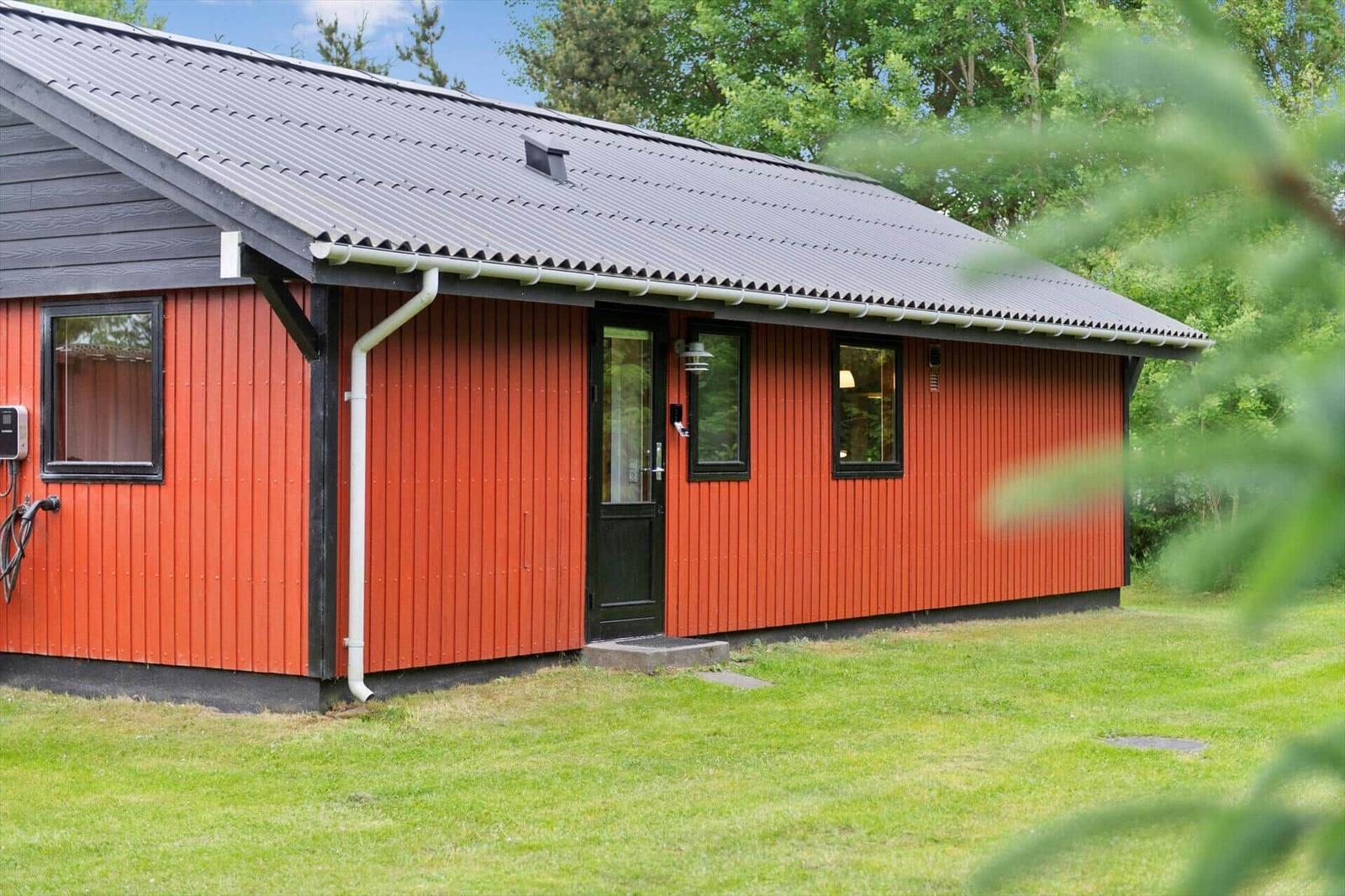 Rotes Holzhaus mit schwarzem Dach und Fenstern. Grasfläche und Bäume im Hintergrund.