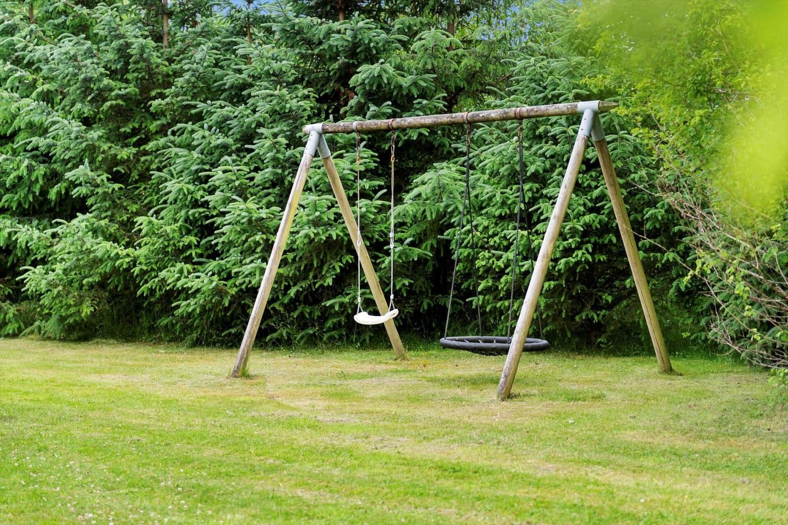 Hinter einem grünen Heckenzaun steht ein Holzschaukelstand mit zwei Schaukeln auf einer Wiese.