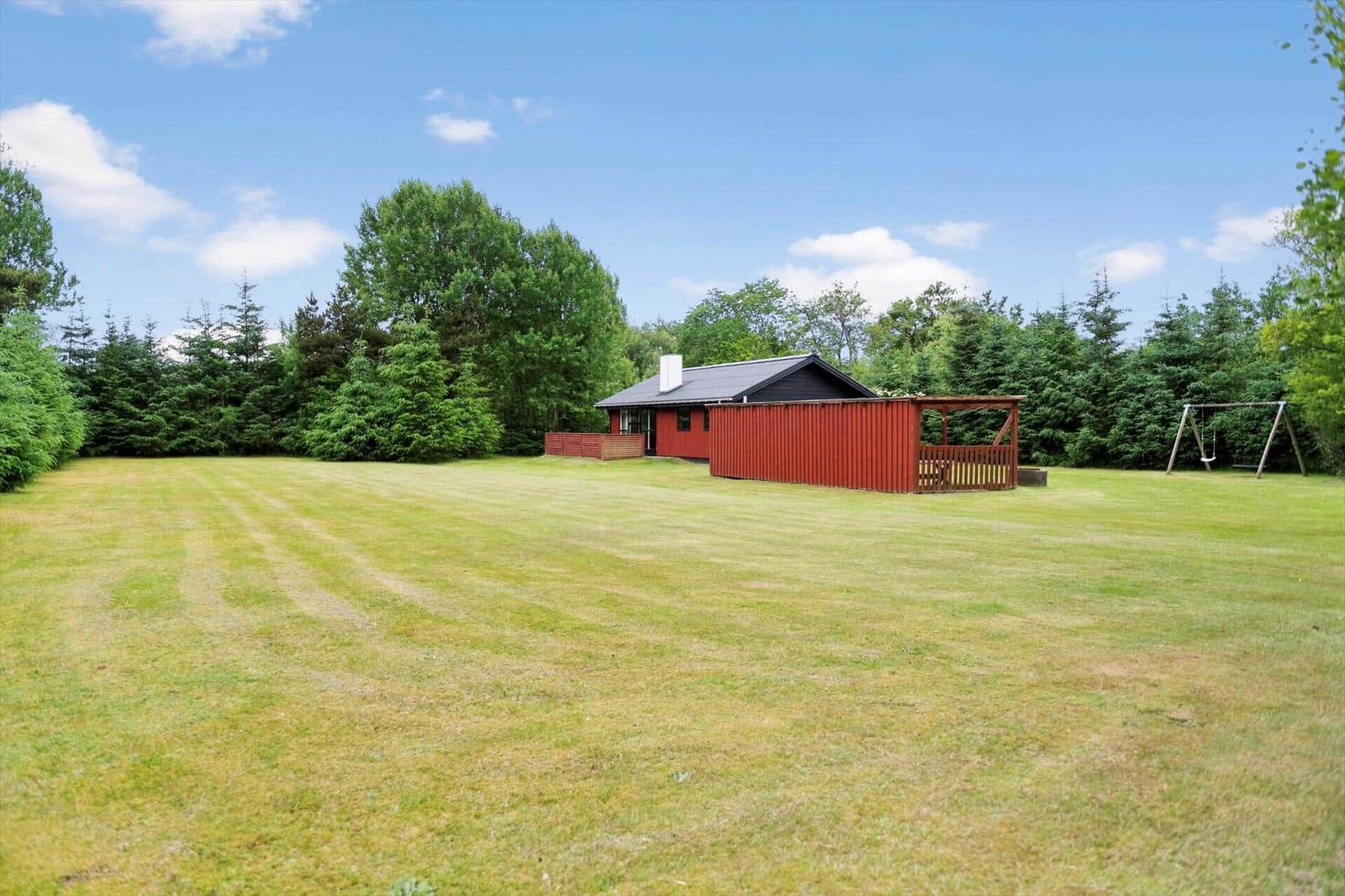 Roter Holzhaus mit Terrasse auf grünem Rasen. Hintergrund: Wald und klarer Himmel.