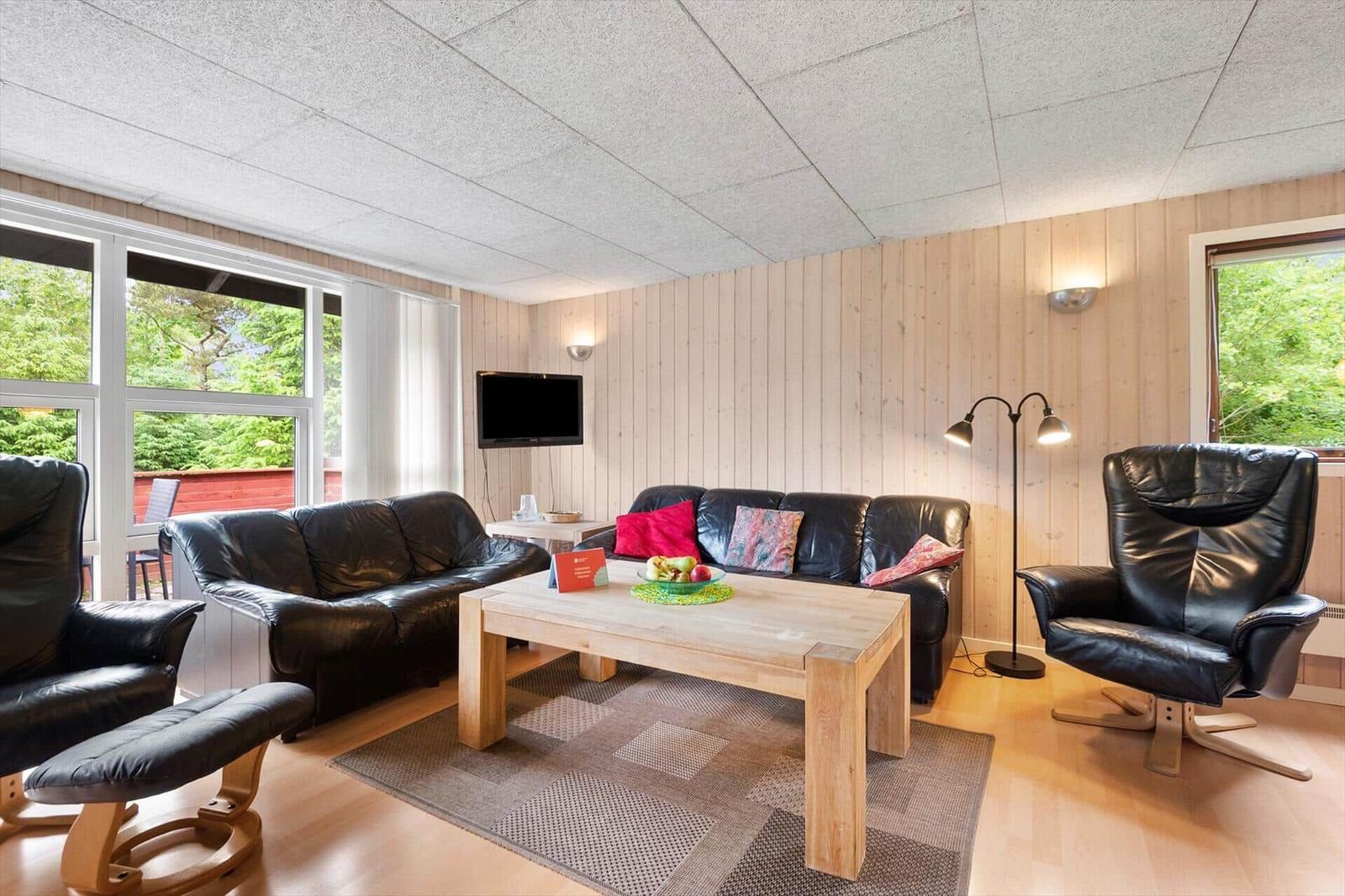Wohnzimmer mit Sofa, Sessel, Tisch und Fernseher. Holzwände und große Fenster.