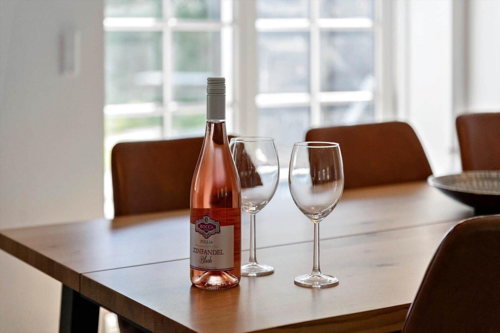 Eine Flasche Roséwein und zwei Gläser stehen auf einem Holztisch. Im Hintergrund sind Stühle und Fenster zu sehen.