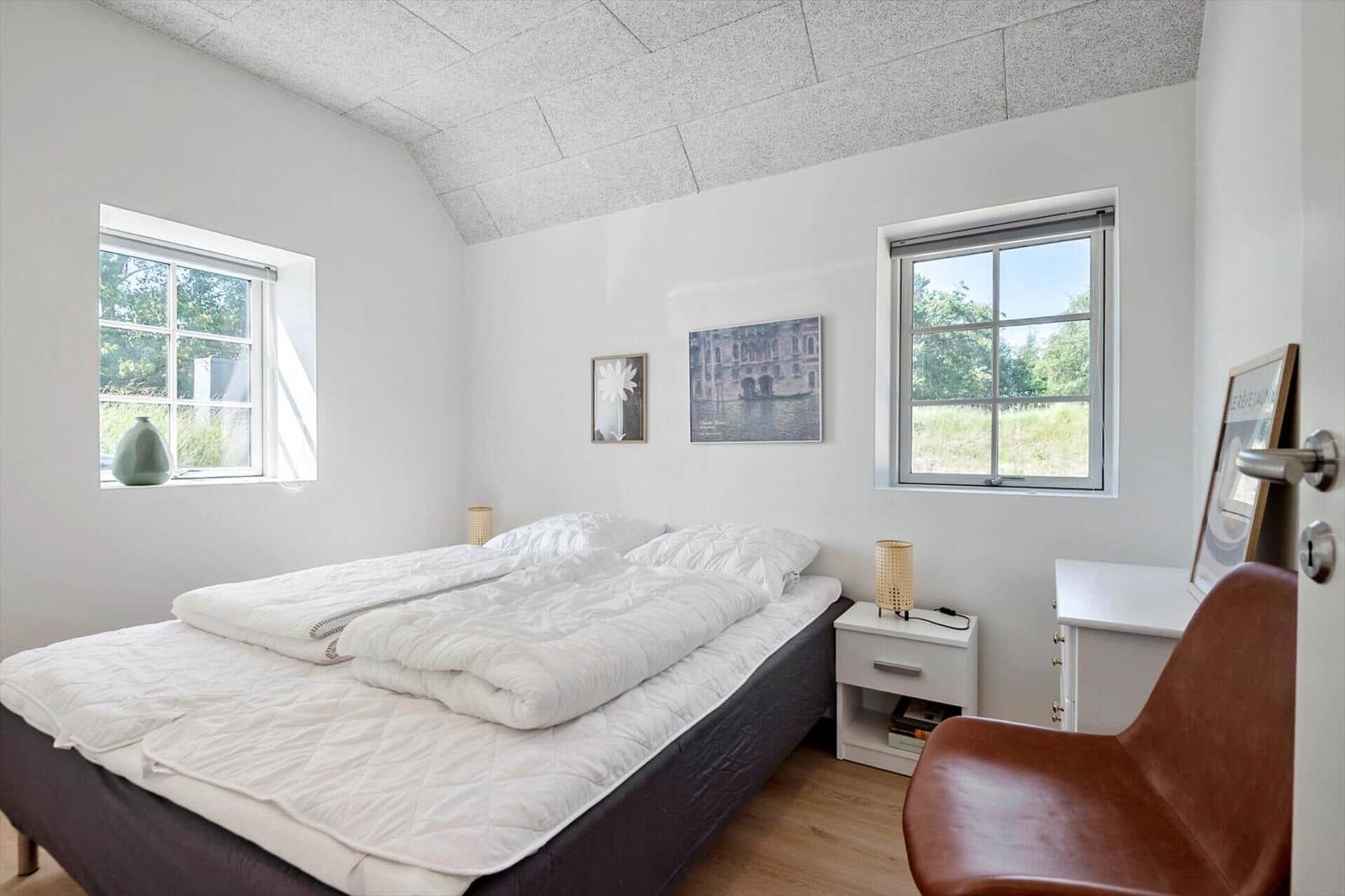 Schlafzimmer mit Bett, zwei Fenstern und einem braunen Stuhl.