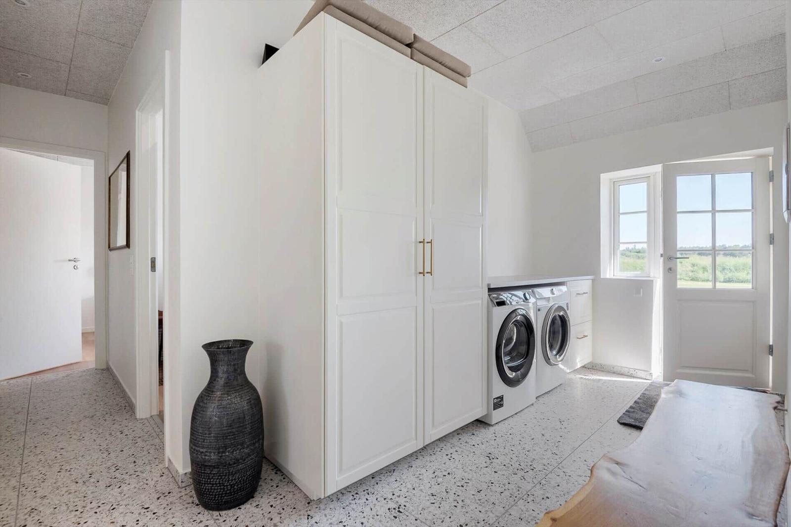 Wäscherei mit Waschmaschine, Trockner und Schrank. Fenster und Tür führen nach draußen.